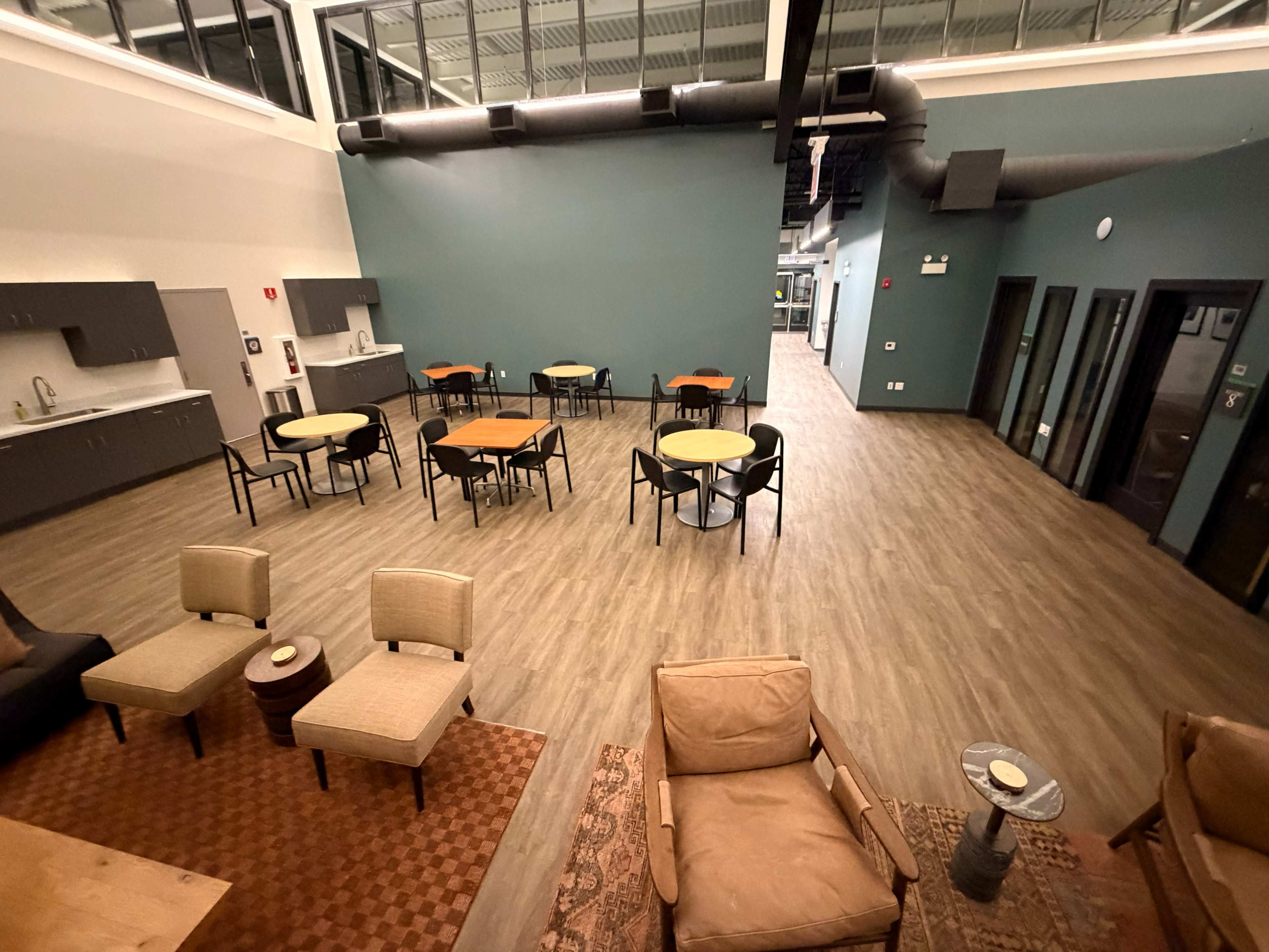 The image shows a spacious and modern interior room with various tables and chairs arranged for seating, along with a kitchenette area in the background.