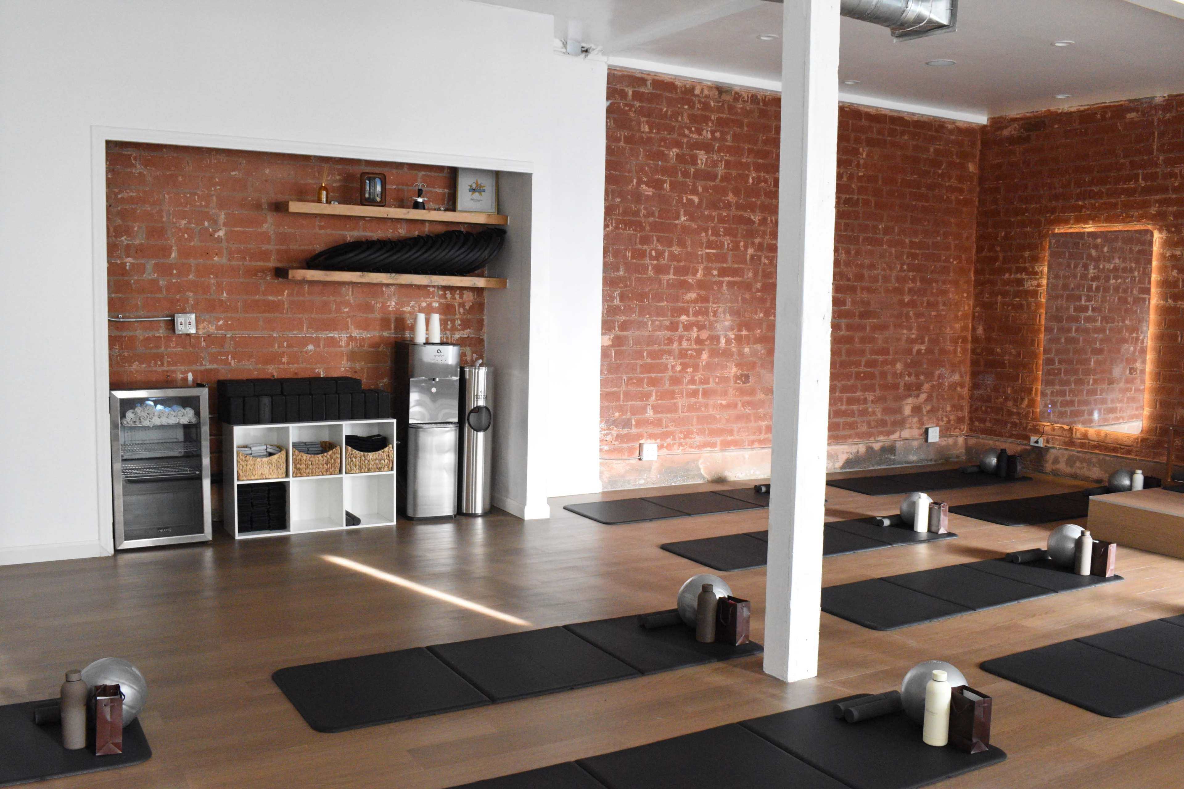 The image shows a yoga studio with dark mats arranged on a wooden floor, a beverage station in the corner, and exposed brick walls.