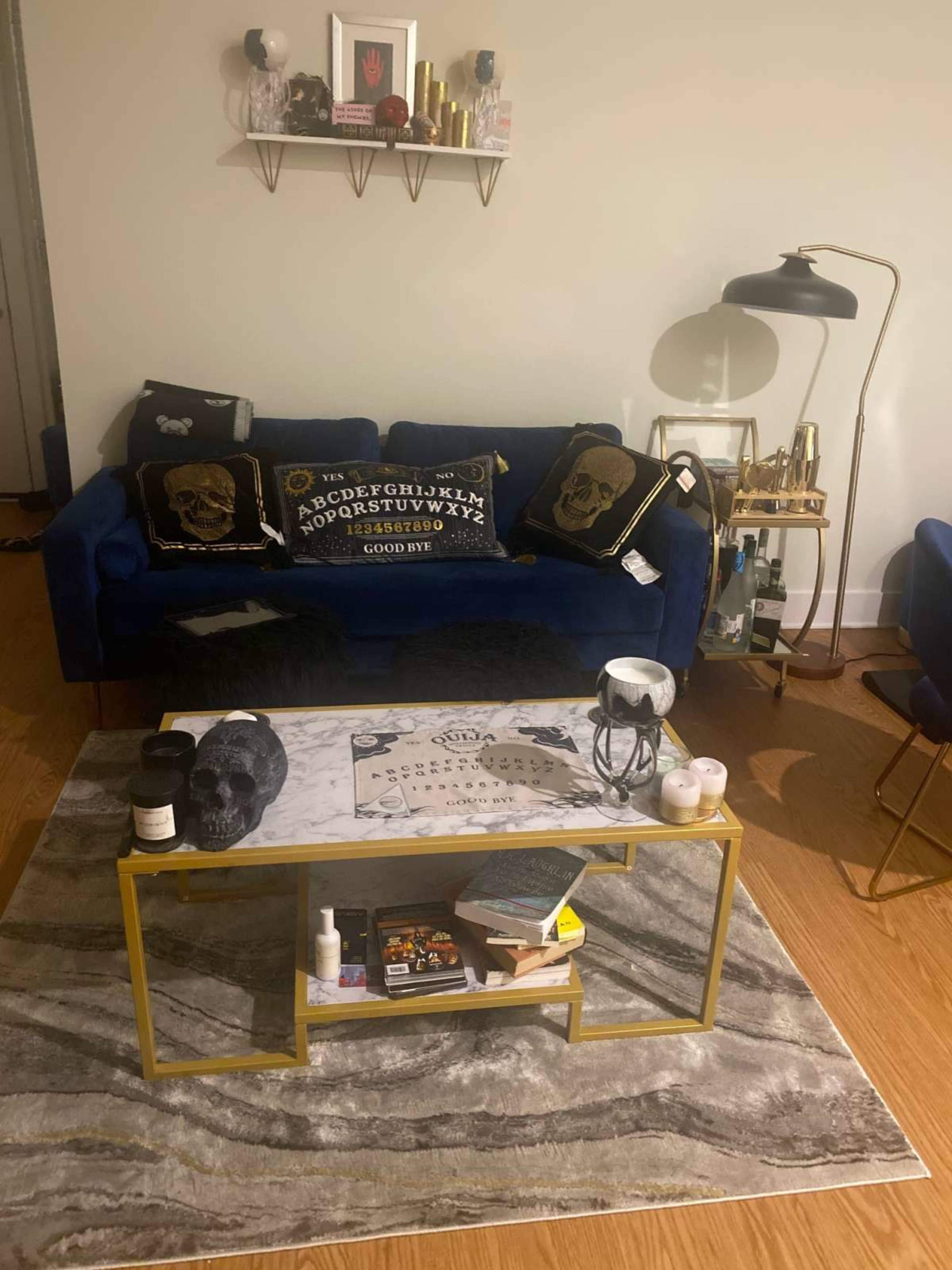 The image shows a living room area with a blue sofa decorated with patterned cushions, a gold coffee table with various items on it, and a floor rug underneath.