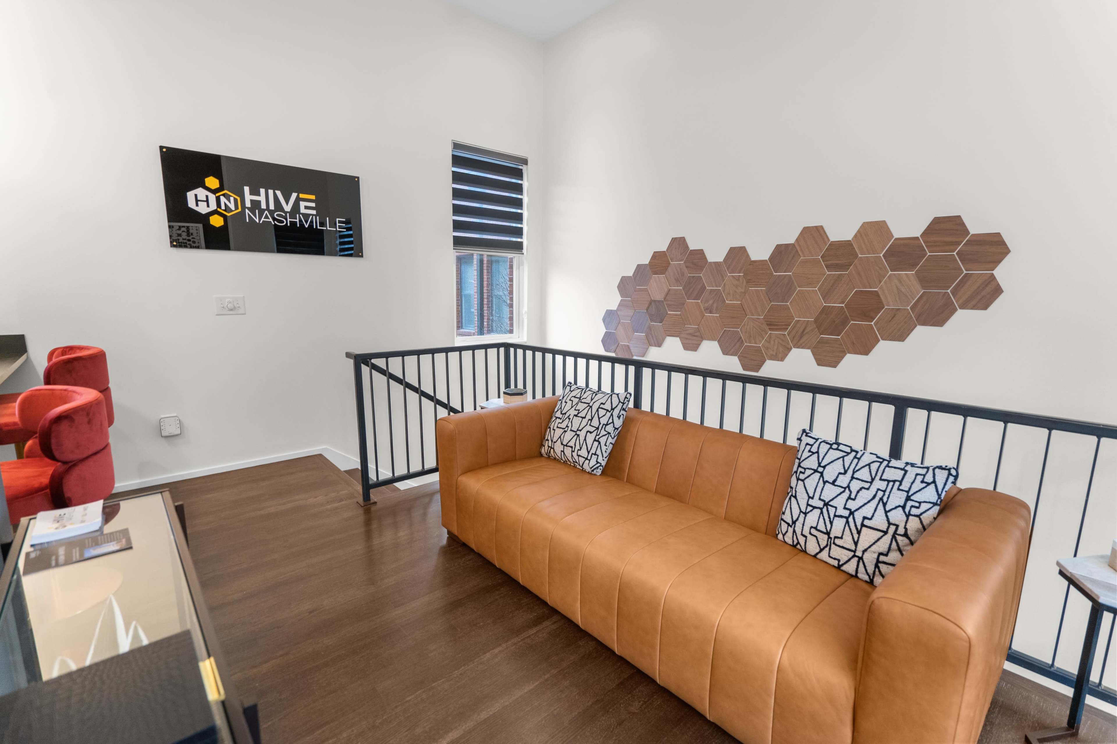 A modern lounge area featuring a tan leather couch with patterned cushions, a black railing, and a decorative hexagonal wall art.