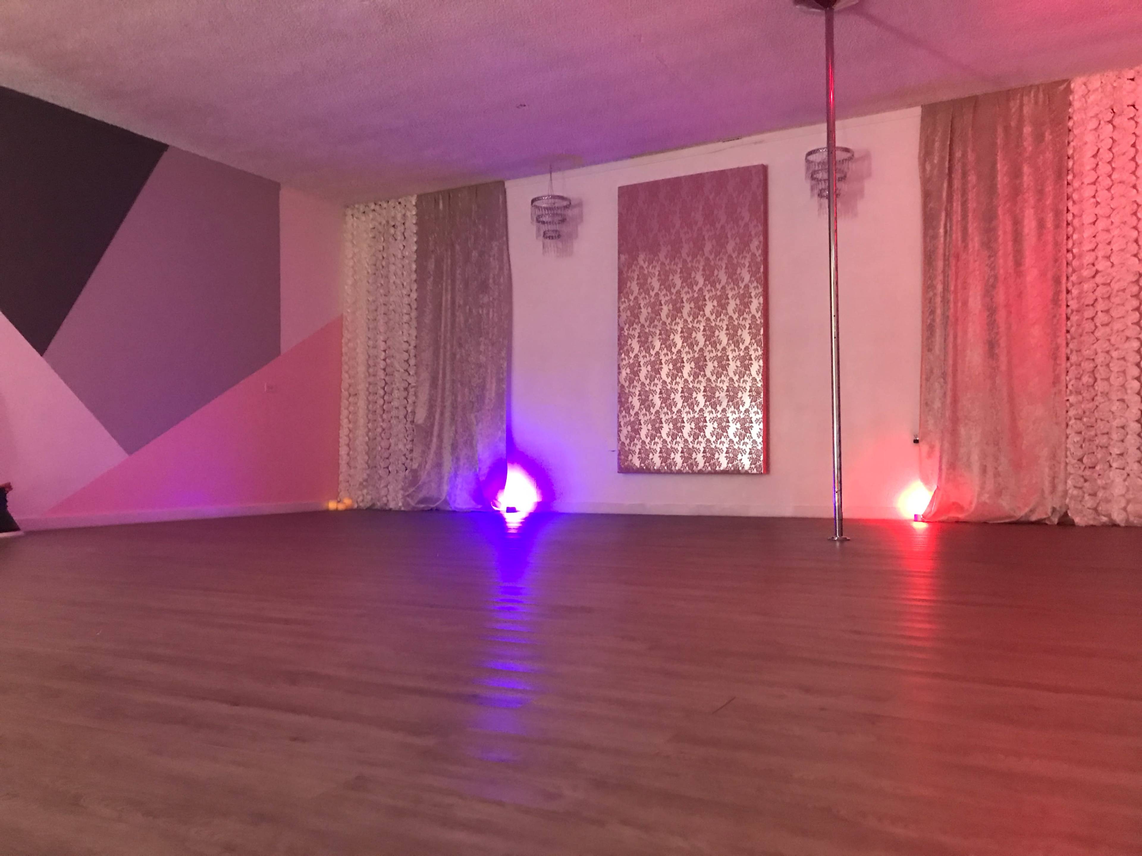 An empty dance studio with a wooden floor, a mirrored wall, decorative curtains, and colored lights at one end.