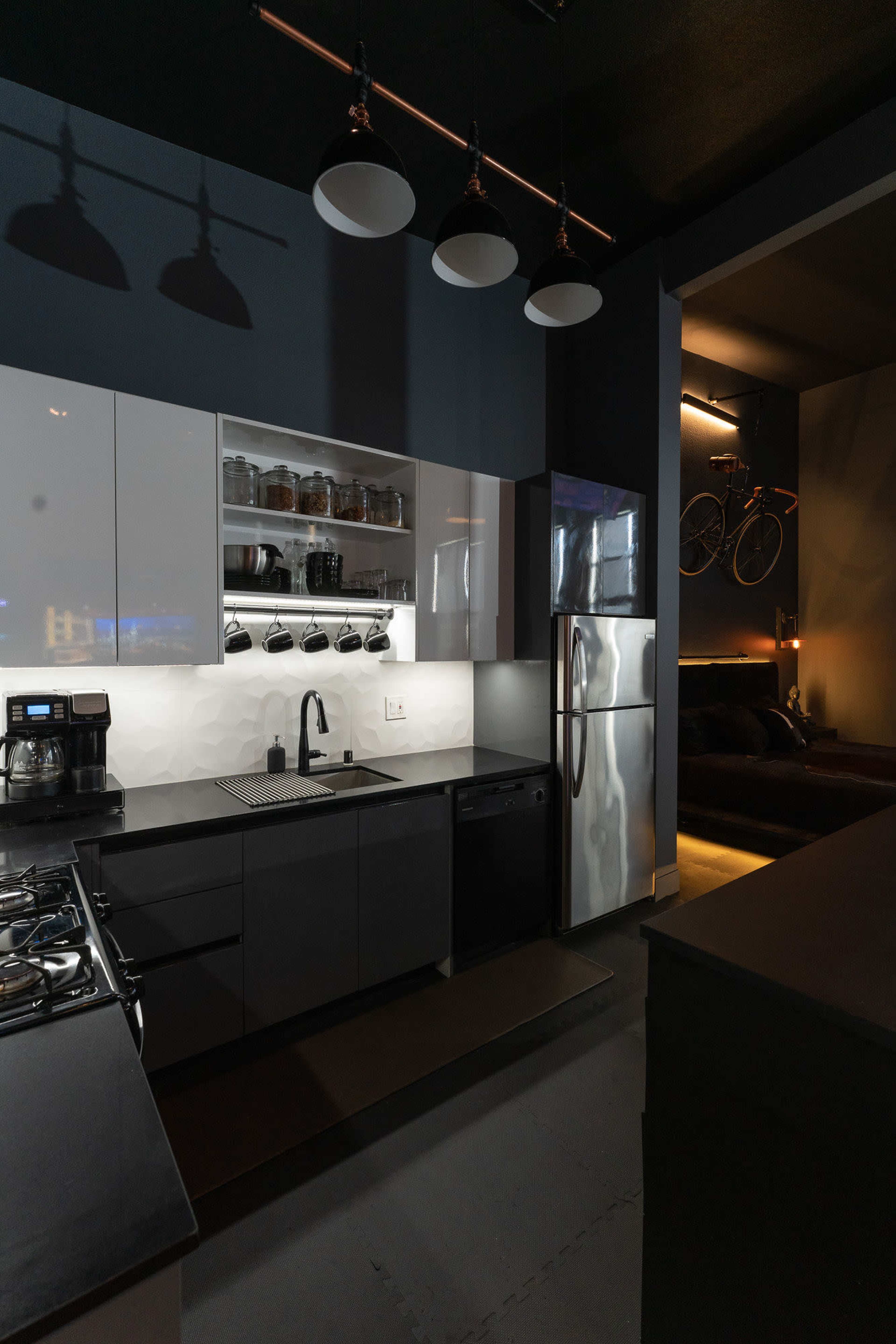 A modern kitchen with dark cabinetry, stainless steel appliances, and minimal lighting, featuring a bike mounted on the wall in an adjacent room.