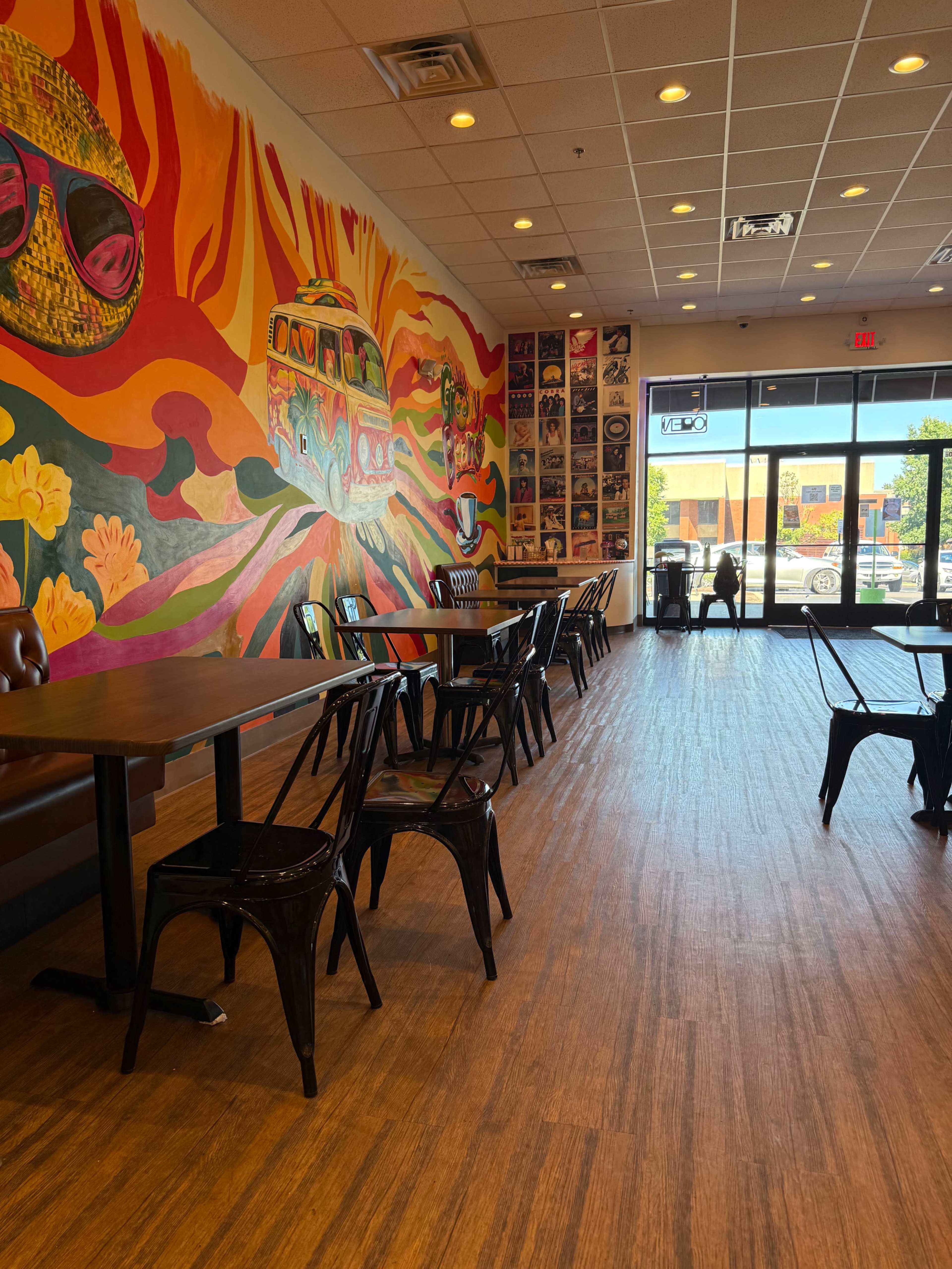 The image shows a brightly colored mural on a cafe wall, with empty tables and chairs arranged inside the establishment, leading to a large window that opens to the outside.