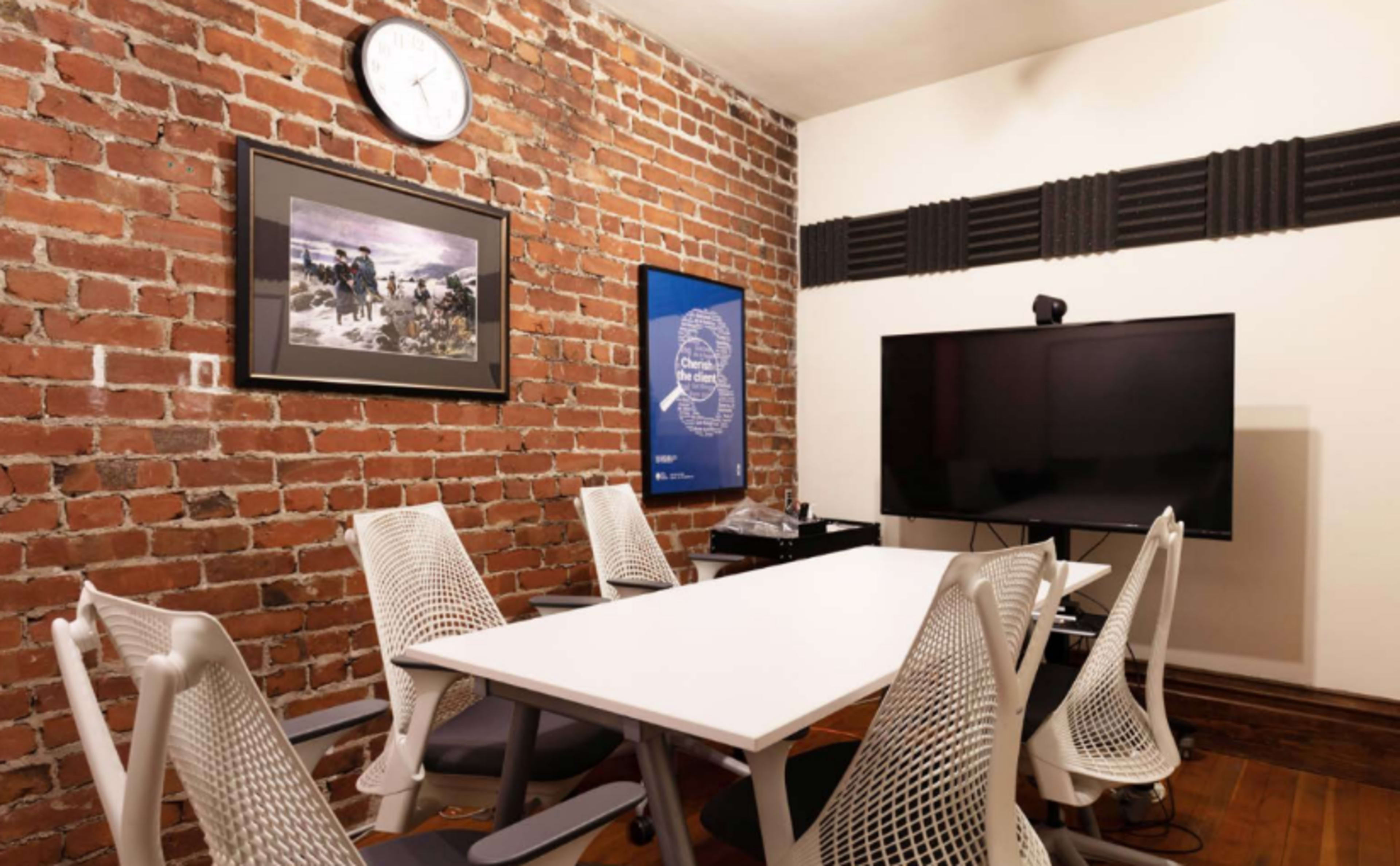 A small meeting room features a white table surrounded by ergonomic chairs, a large television, and exposed brick walls adorned with framed artwork.