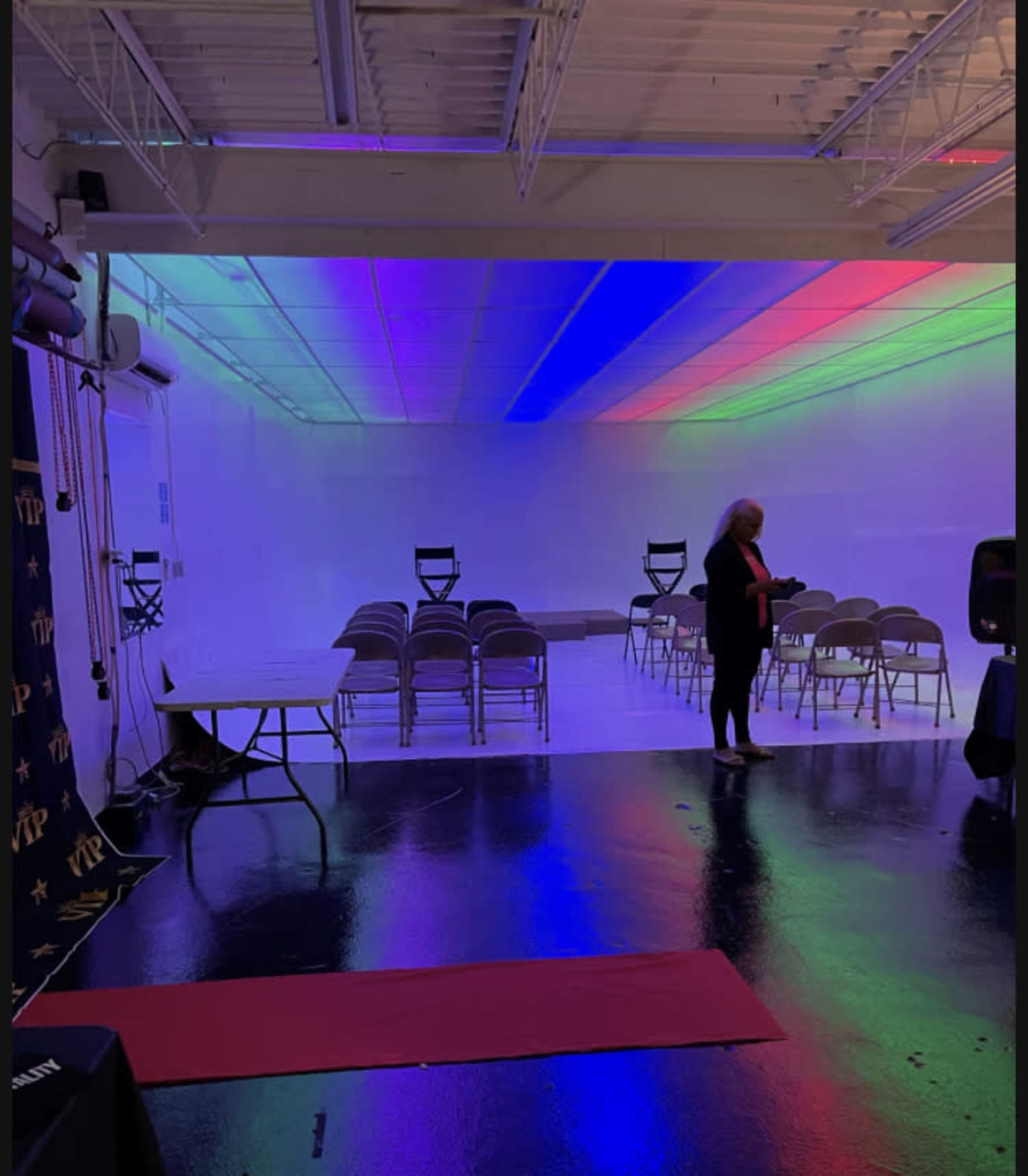 A brightly lit room features a row of chairs, a table, and a woman standing near a red carpet under colorful lighting.