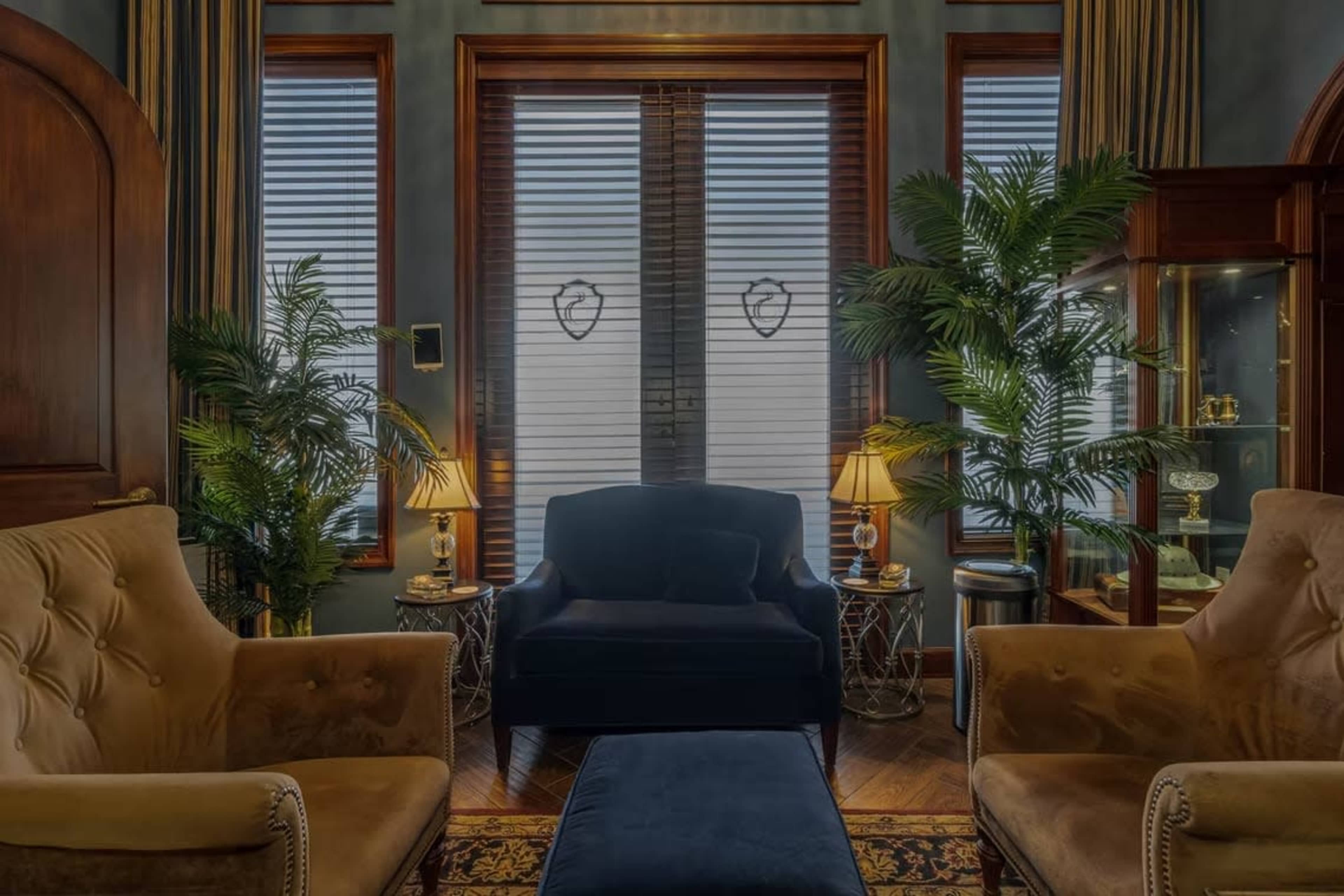 The image shows a cozy sitting area with two armchairs, a central ottoman, and decorative plants, illuminated by table lamps near the windows.