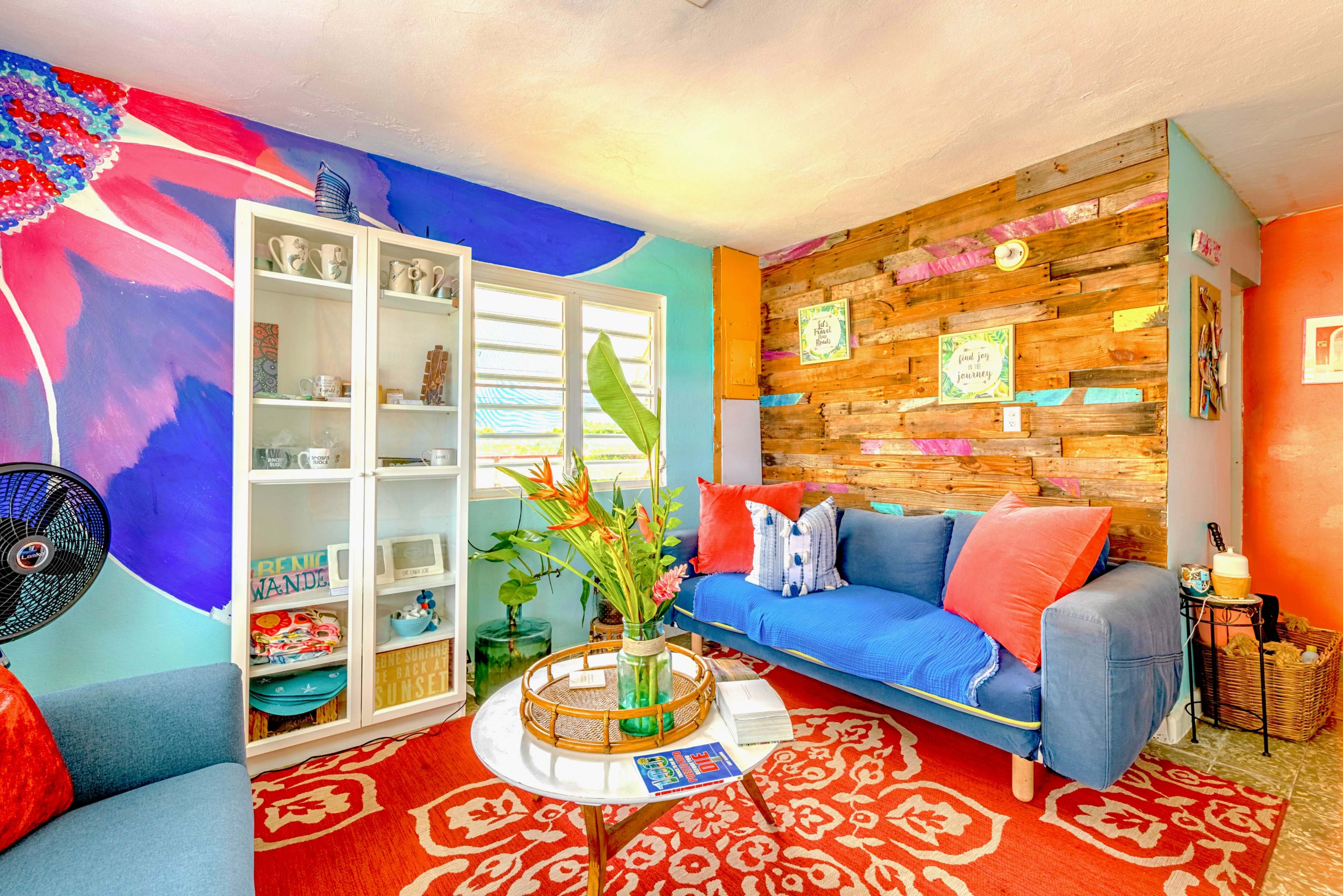The image shows a colorful living room featuring a blue sofa, a wooden accent wall, a glass display cabinet, and vibrant decorative elements.