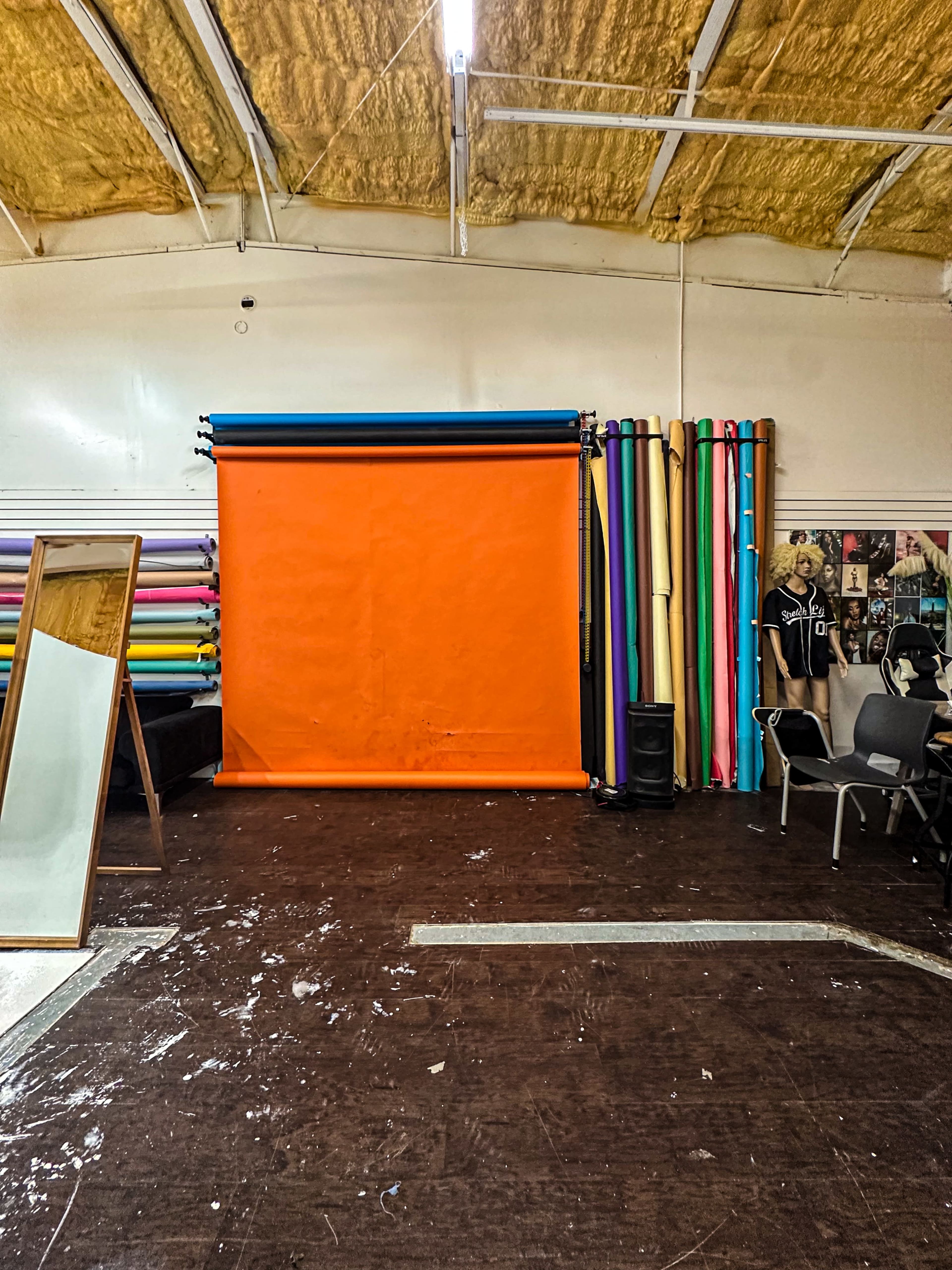 The image shows a studio space with an orange backdrop, various rolls of colored paper along the walls, and a large mirror.