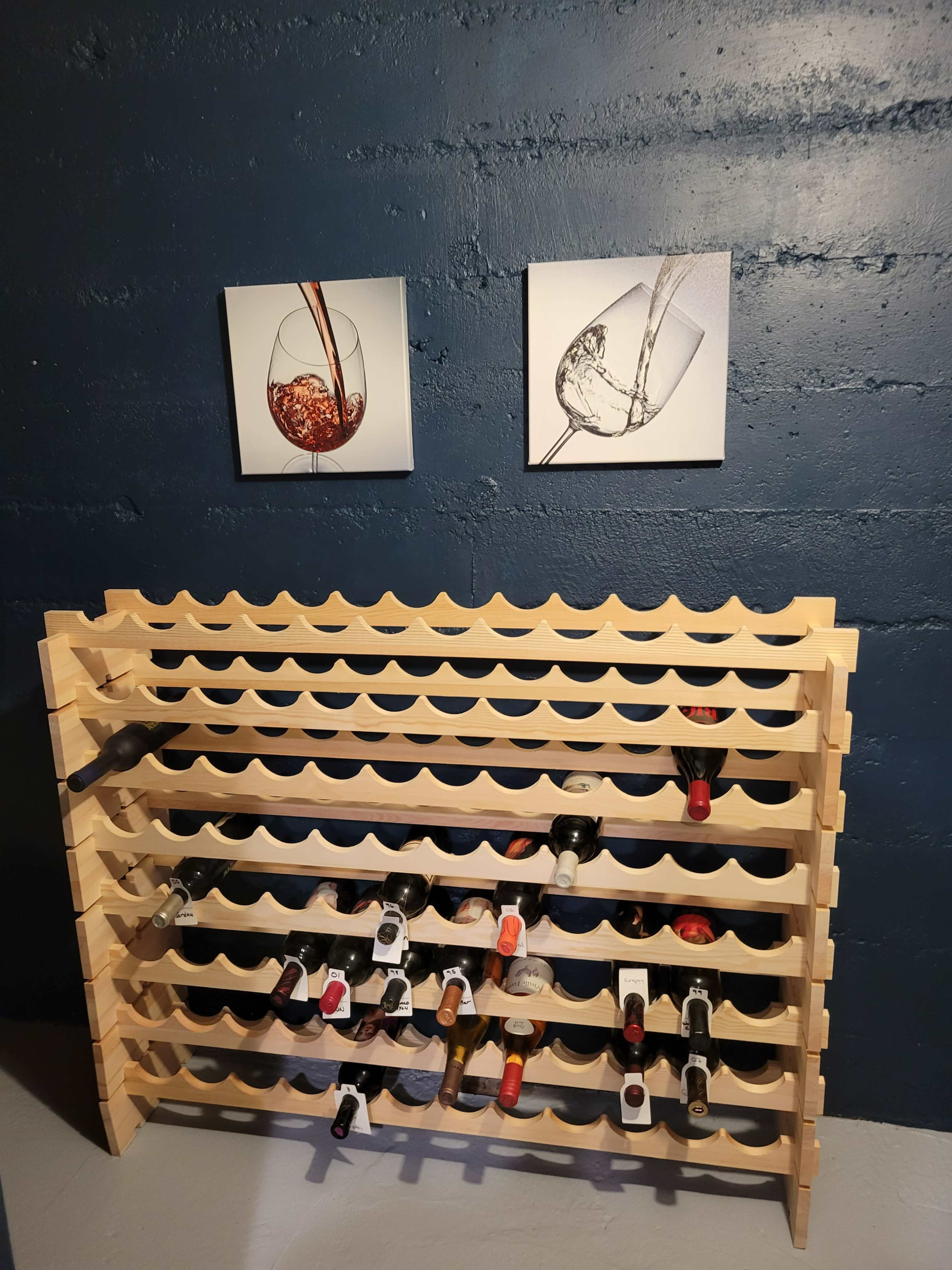 A wooden wine rack holding several bottles is positioned against a dark blue wall, above which are two framed images of wine glasses.
