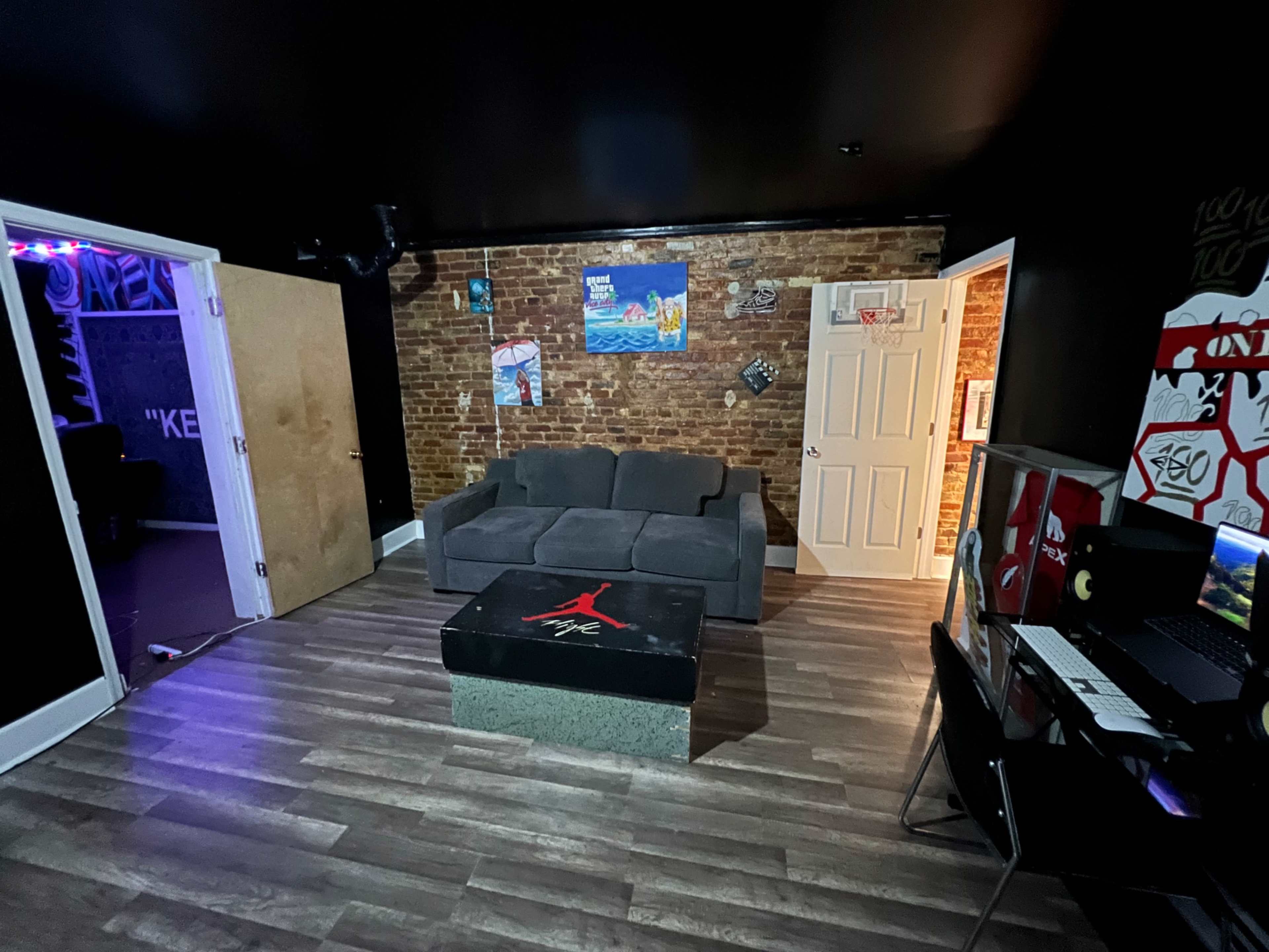 A room with a gray couch, a coffee table featuring a black Jordan logo, and a desk with a computer, all set against exposed brick walls and dark paint.