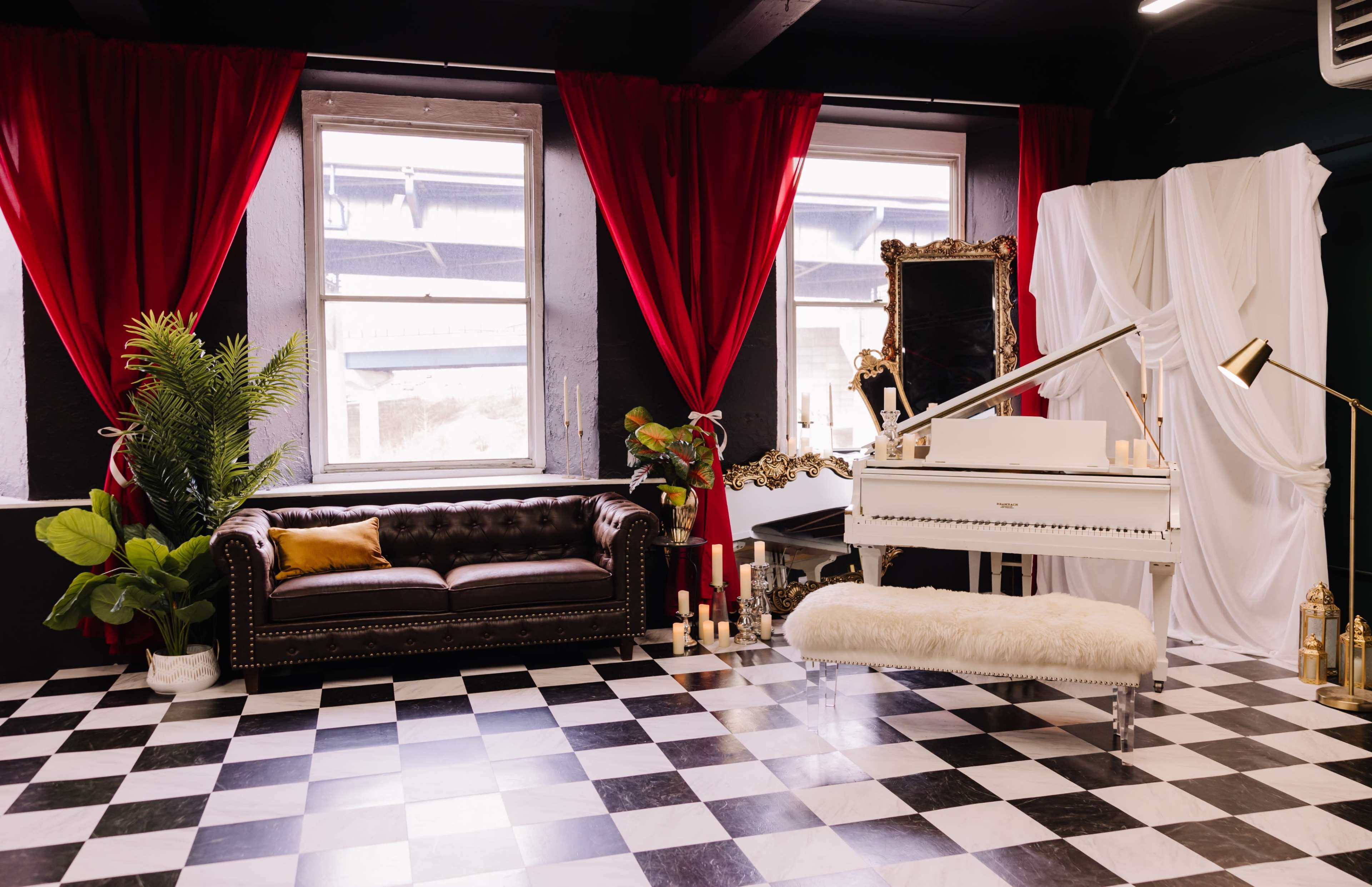 The scene features a stylish interior with a black and white checkered floor, a brown leather sofa, a white piano, and decorative plants, accentuated by red curtains and soft lighting.