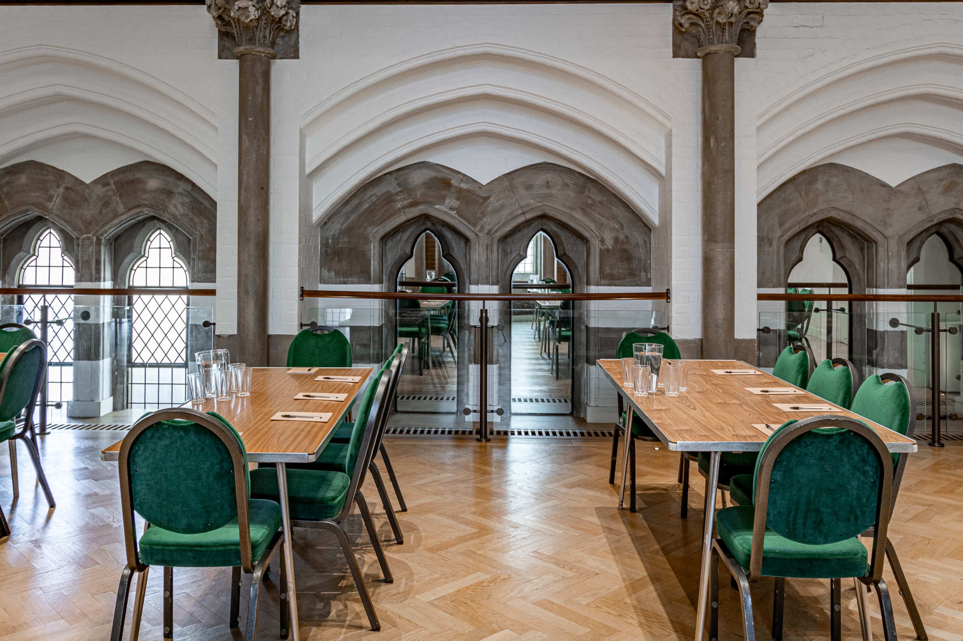 The image shows a dining area with several tables set for meals, featuring green chairs and large arched windows in the background.