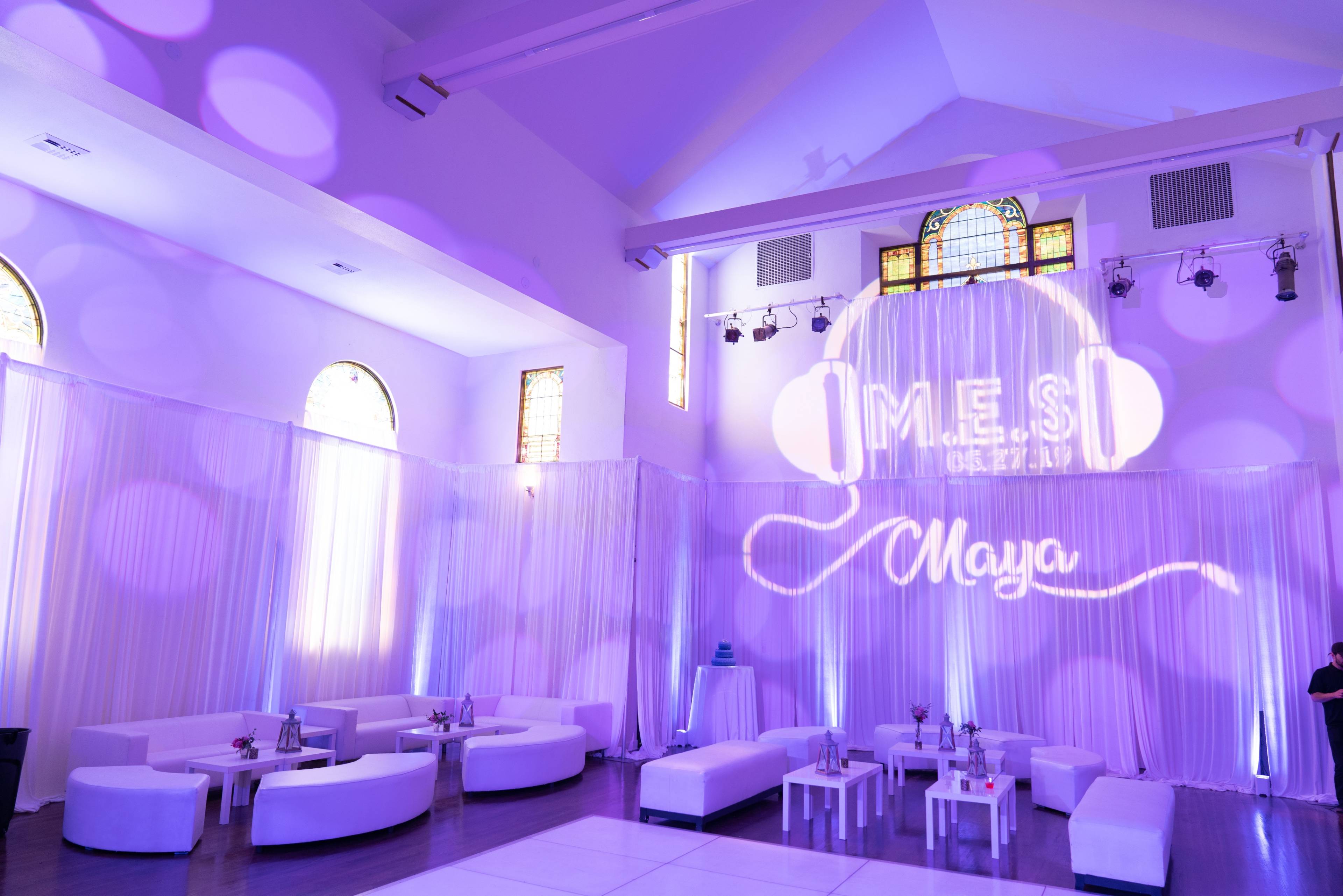 The image shows a well-decorated event space with white lounge furniture, purple lighting, and a large projection featuring the name "Maya" and headphones on the wall.