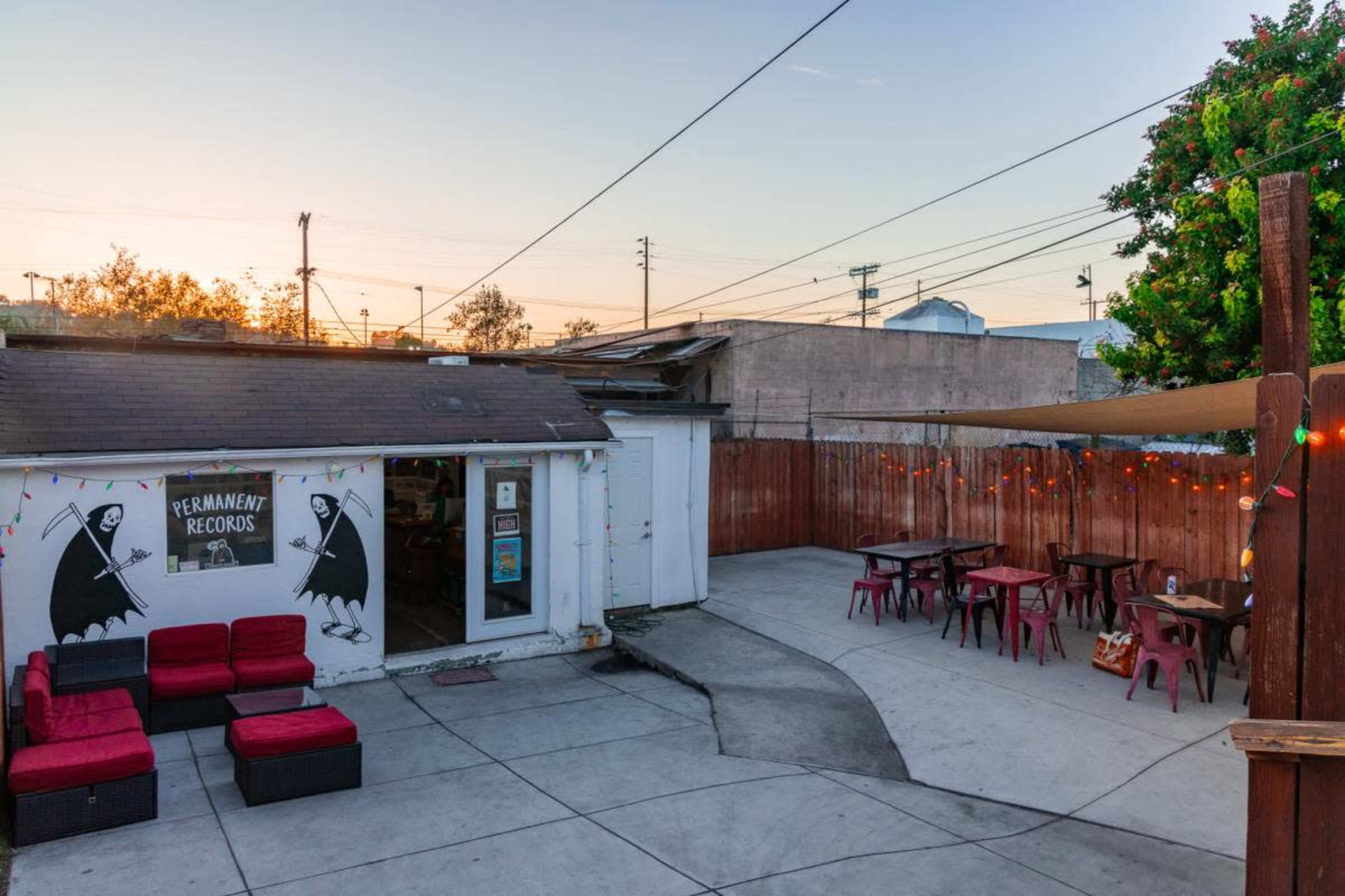 The image shows a small outdoor area of a record store, featuring a seating arrangement with red chairs and tables, alongside a wall adorned with murals.