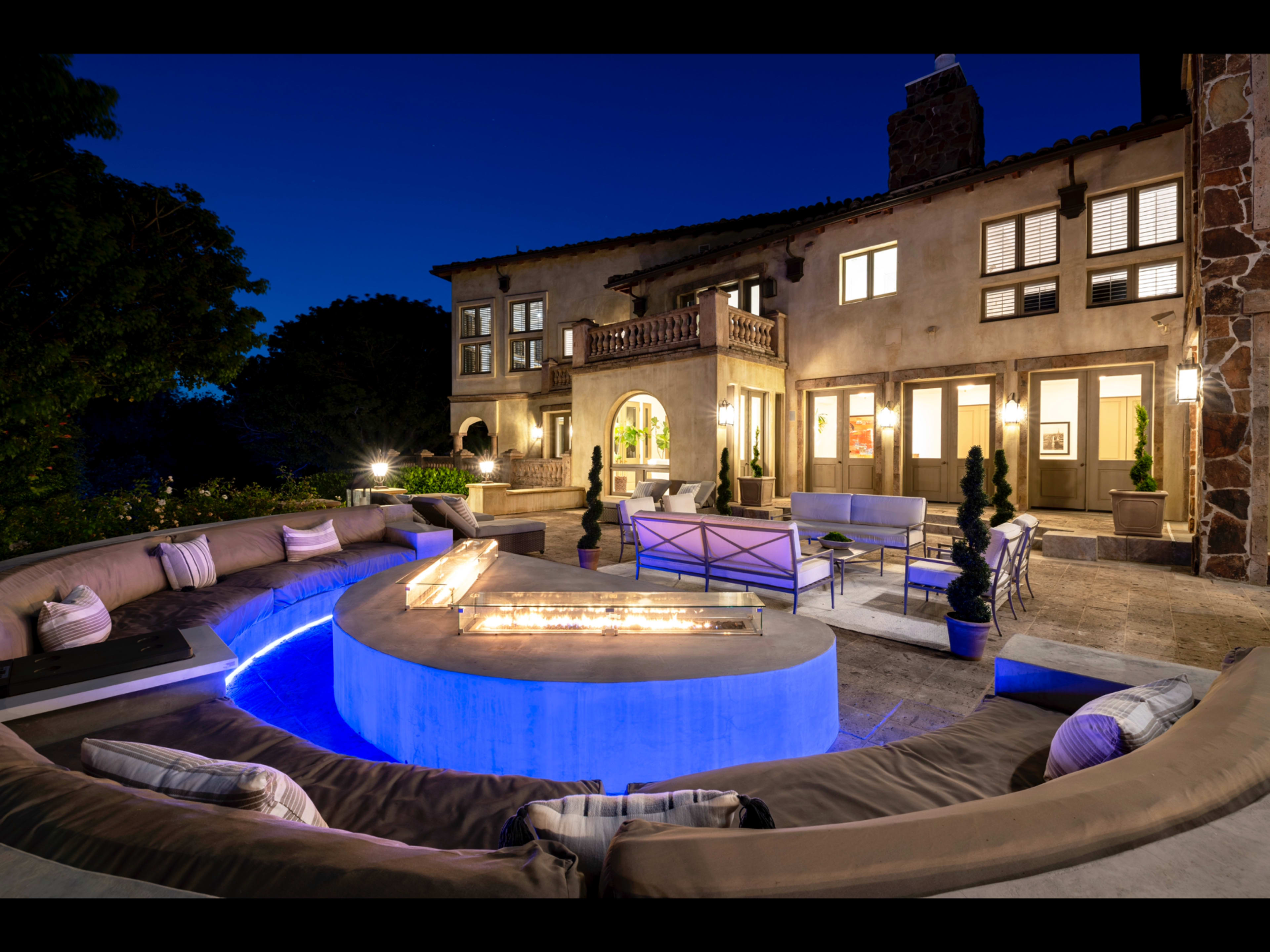 The image shows a modern outdoor living space with a circular fire pit surrounded by a curved bench, set against a backdrop of a well-lit multi-story home at dusk.