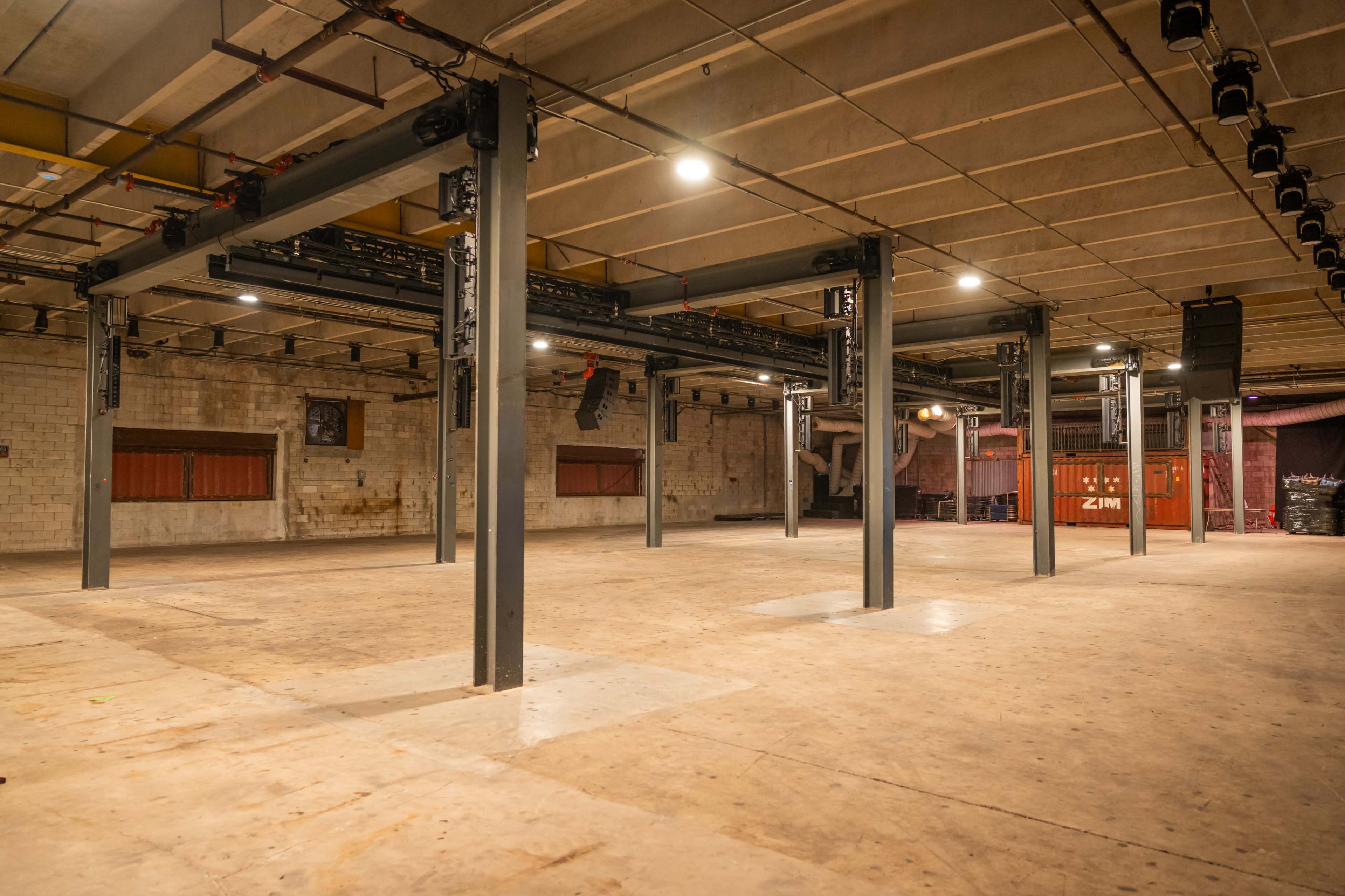 The image shows an empty industrial-style venue with exposed concrete walls, overhead lighting, and metal support beams.