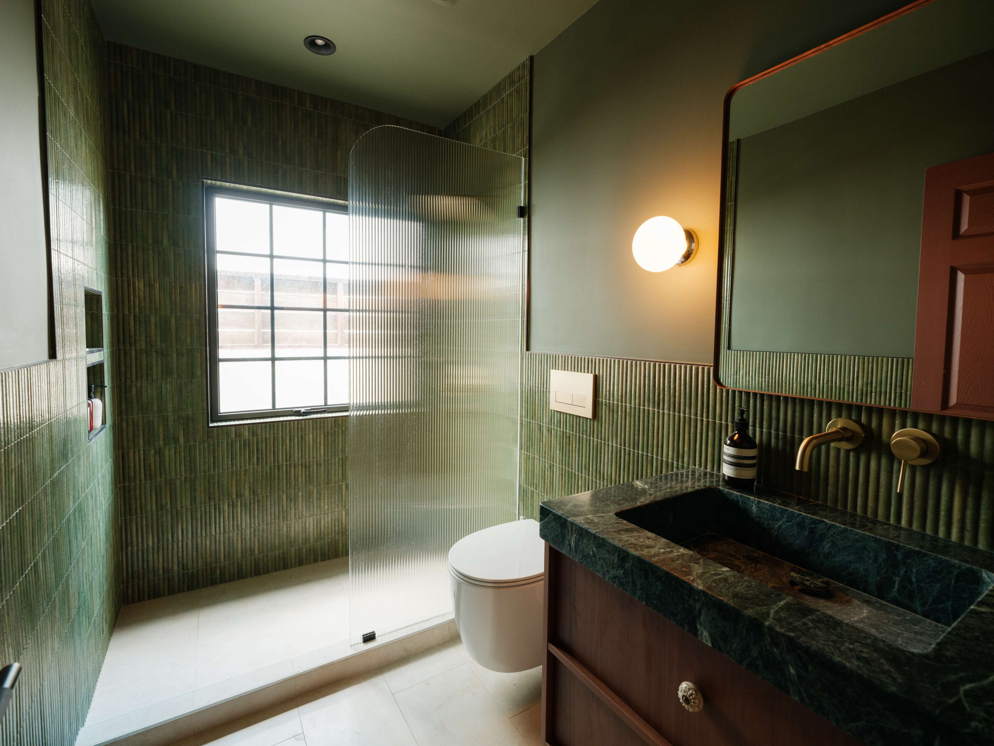 The image shows a modern bathroom featuring green tiled walls, a glass shower enclosure, a toilet, and a marble countertop with a sink.