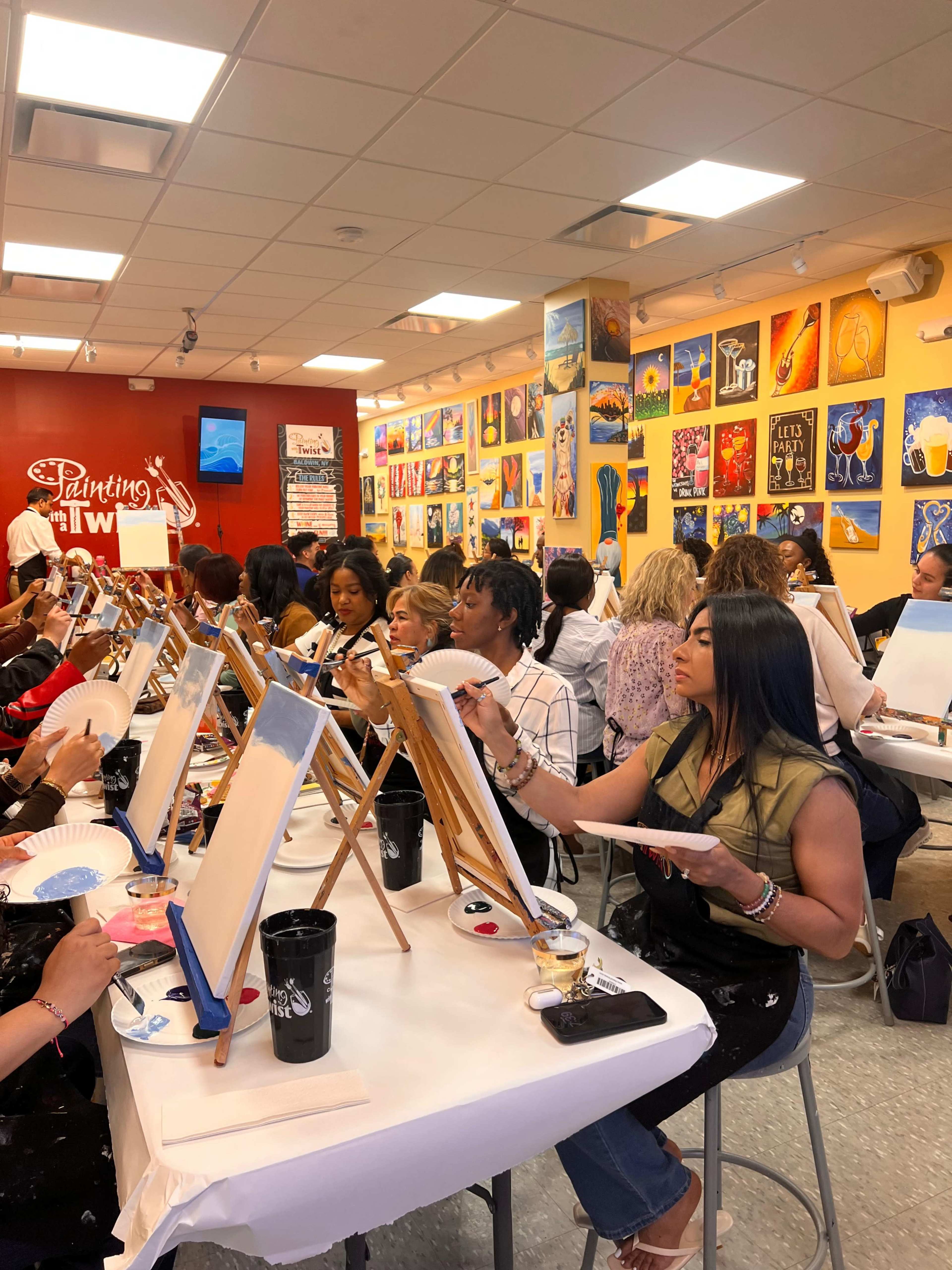 A group of people is sitting at tables in a studio, each working on canvas paintings while surrounded by colorful artwork on the walls.