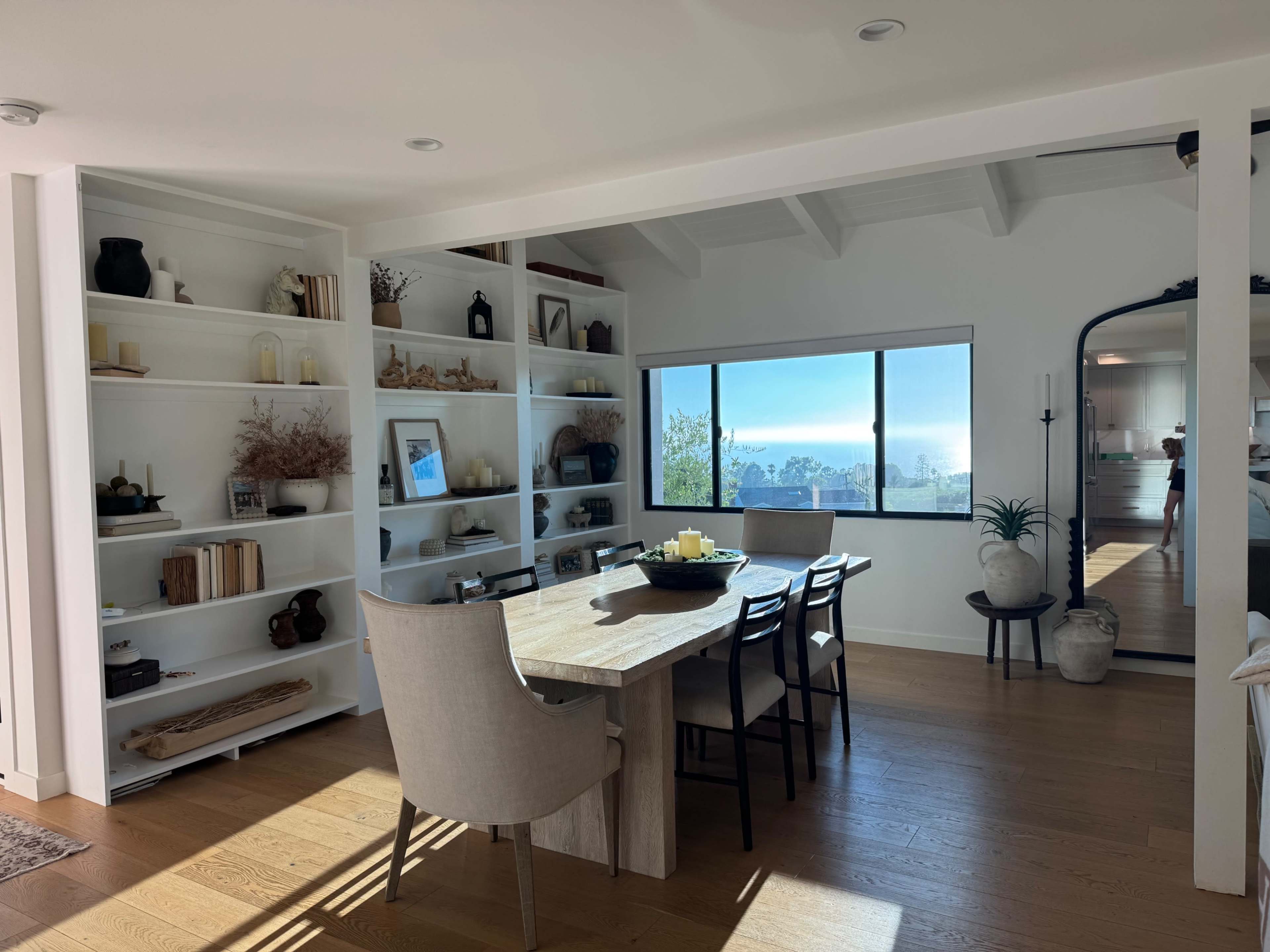 A bright dining area features a wooden table surrounded by black chairs, with shelves displaying various decorative items and a large window offering a scenic view.