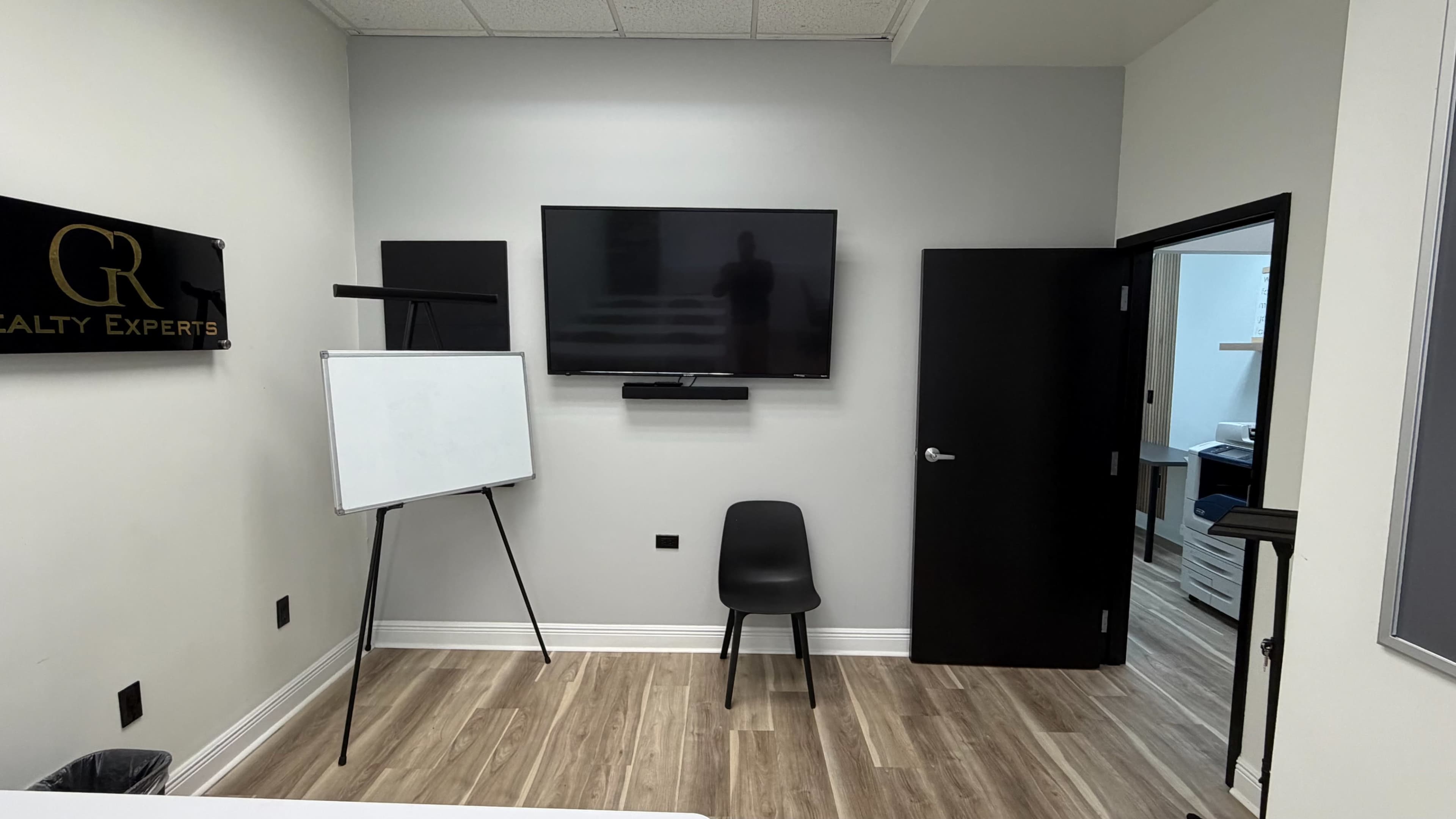 The image shows a modern office space with a wall-mounted television, a whiteboard on an easel, a single black chair, and a doorway leading to another room.