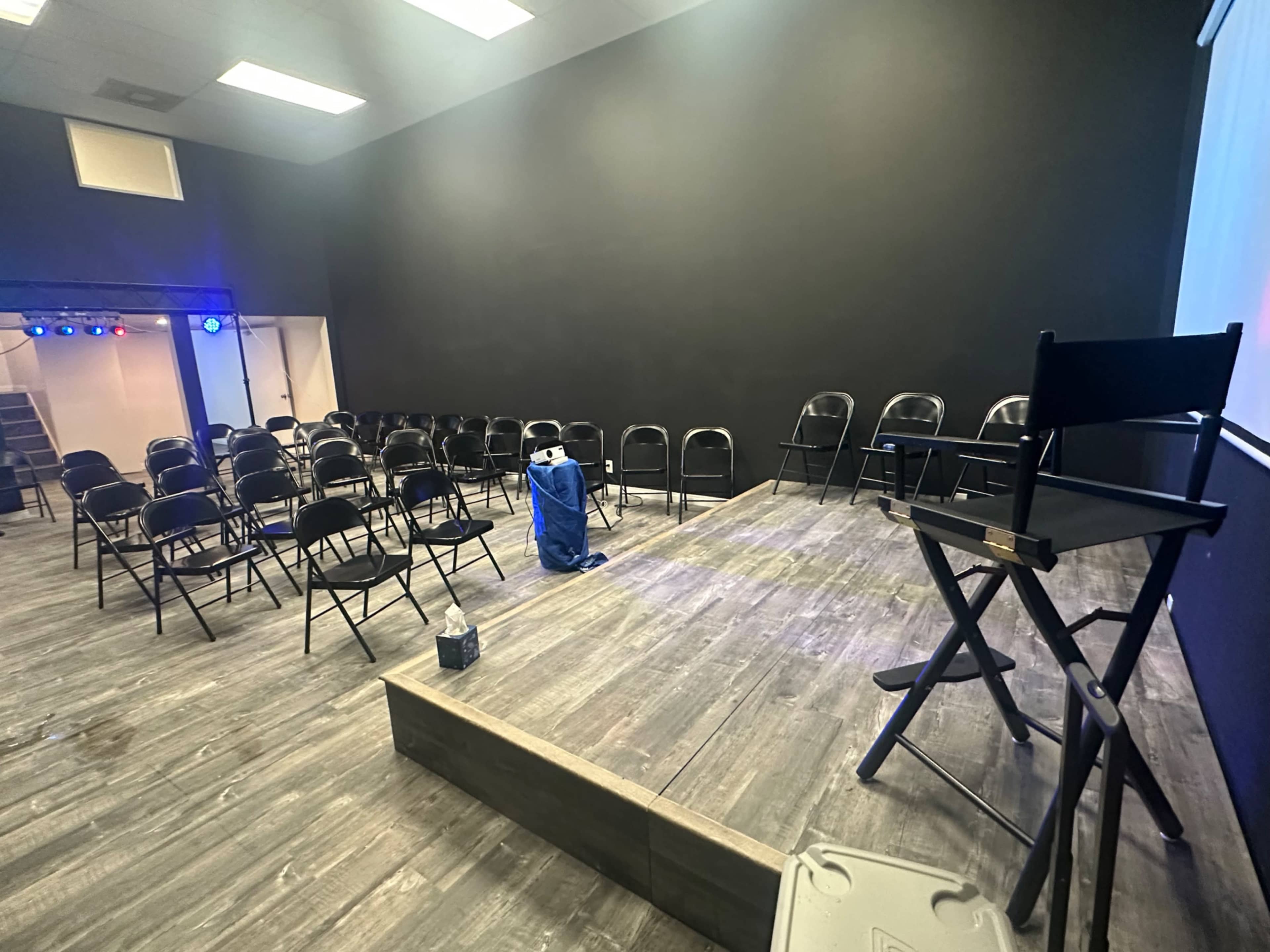 The image shows a small stage area with a row of black chairs arranged for an audience and a director's chair placed on an elevated platform.