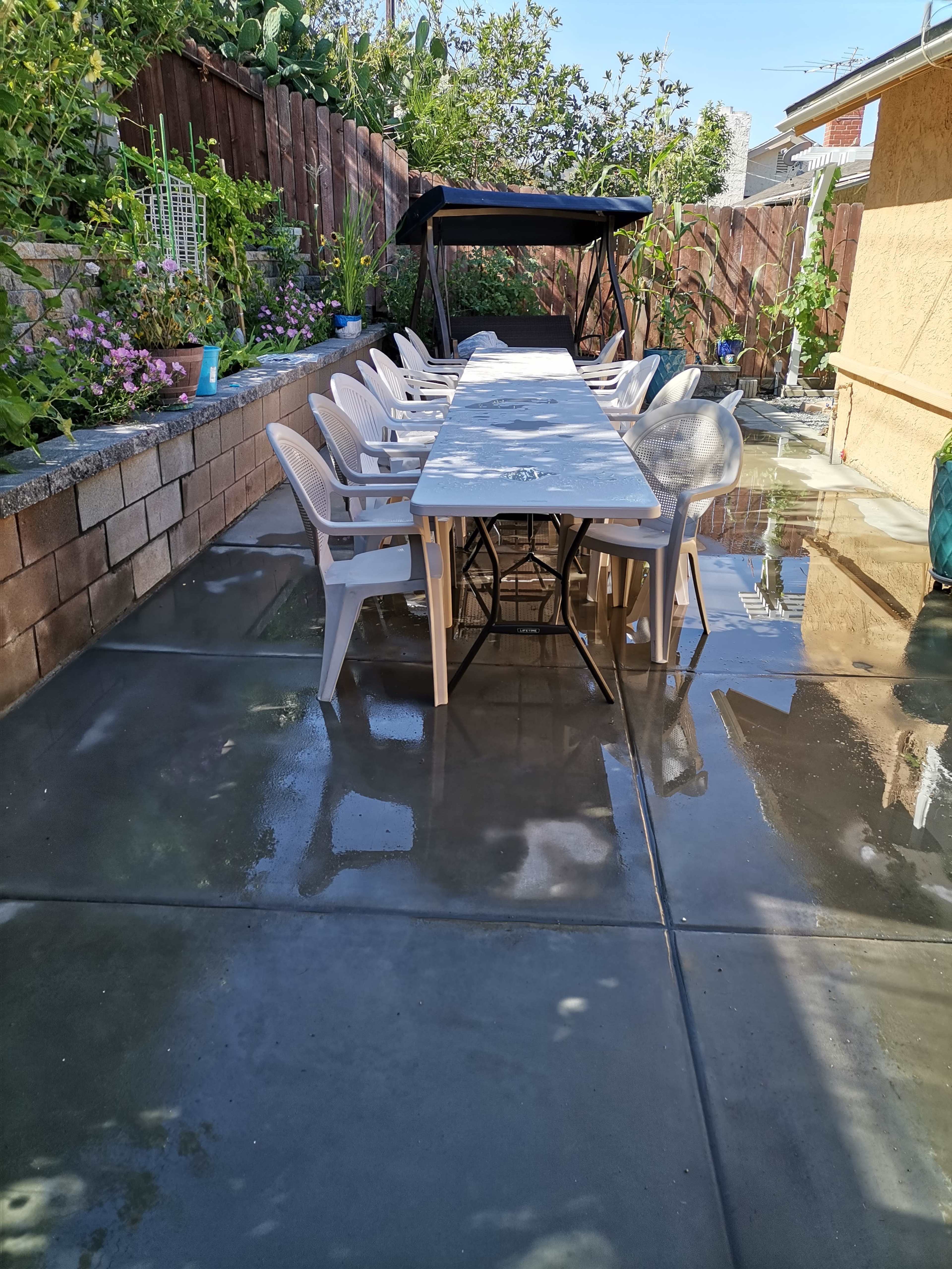 A long table with chairs is set up on a wet concrete patio surrounded by greenery and a shaded seating area.