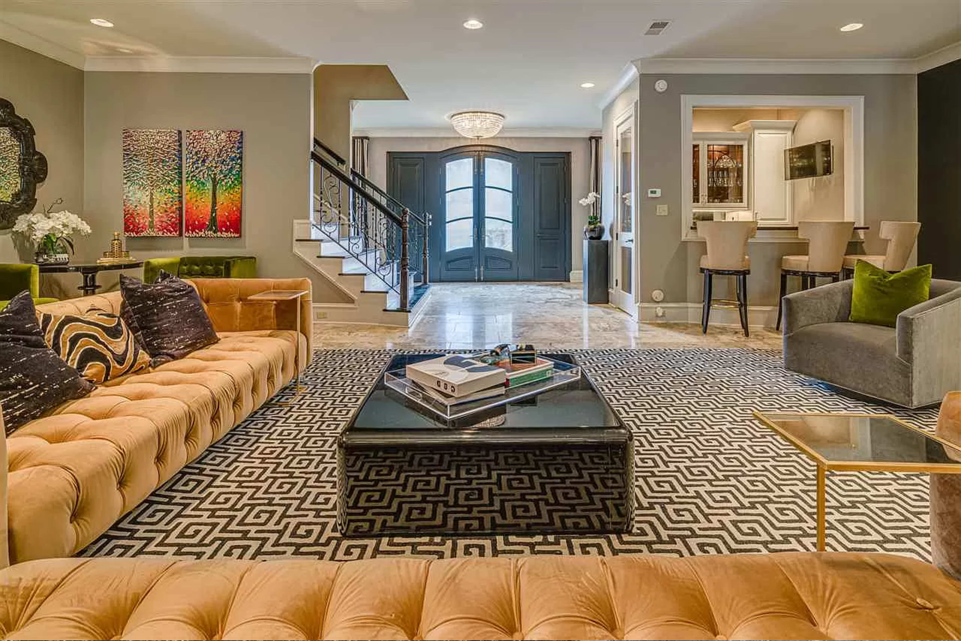A spacious living room featuring a large tufted sofa, a rectangular coffee table, and a patterned area rug, with elegant decor and a staircase in the background.
