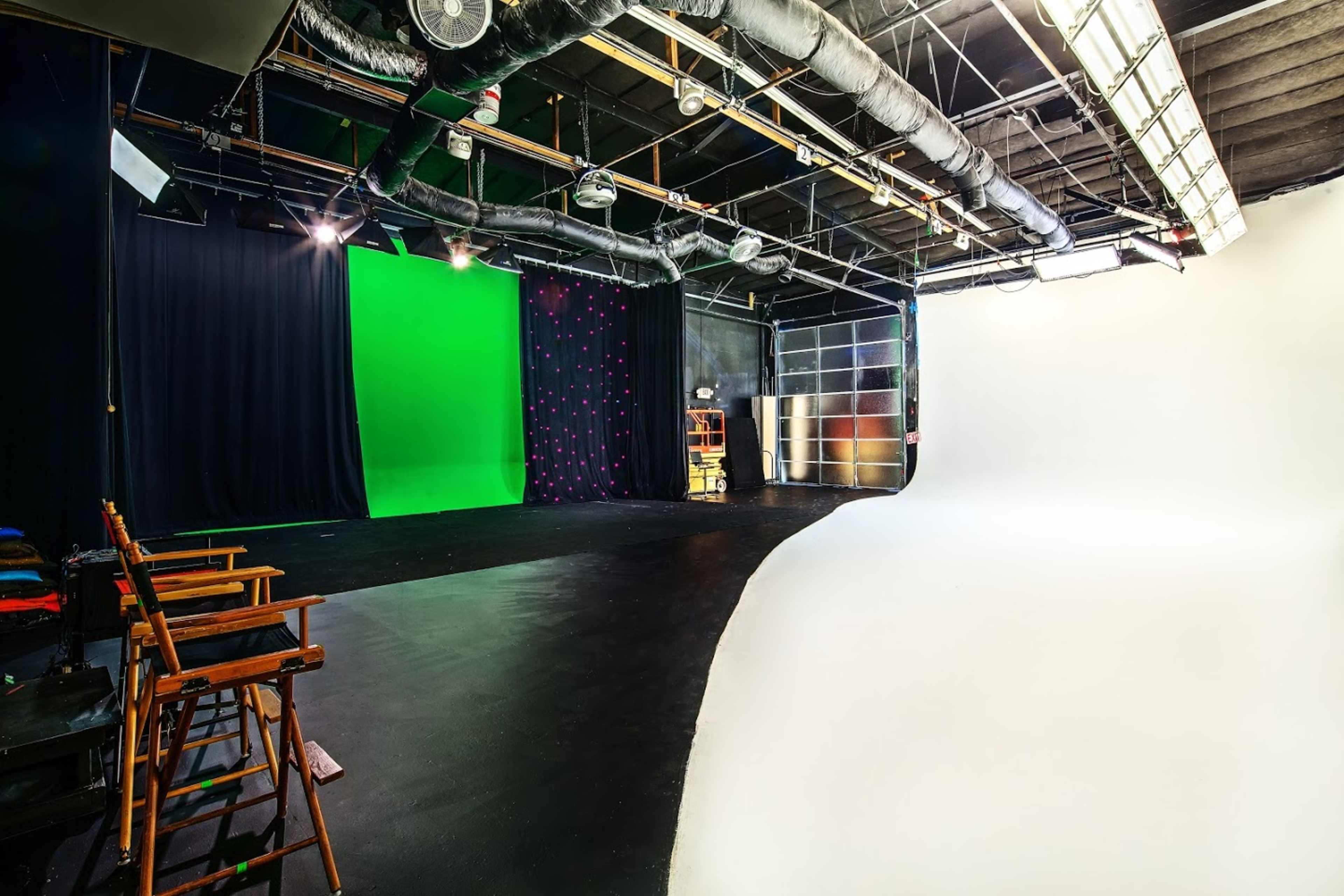 A sparsely furnished film studio with a green screen backdrop on one side and a curved white backdrop on the other.