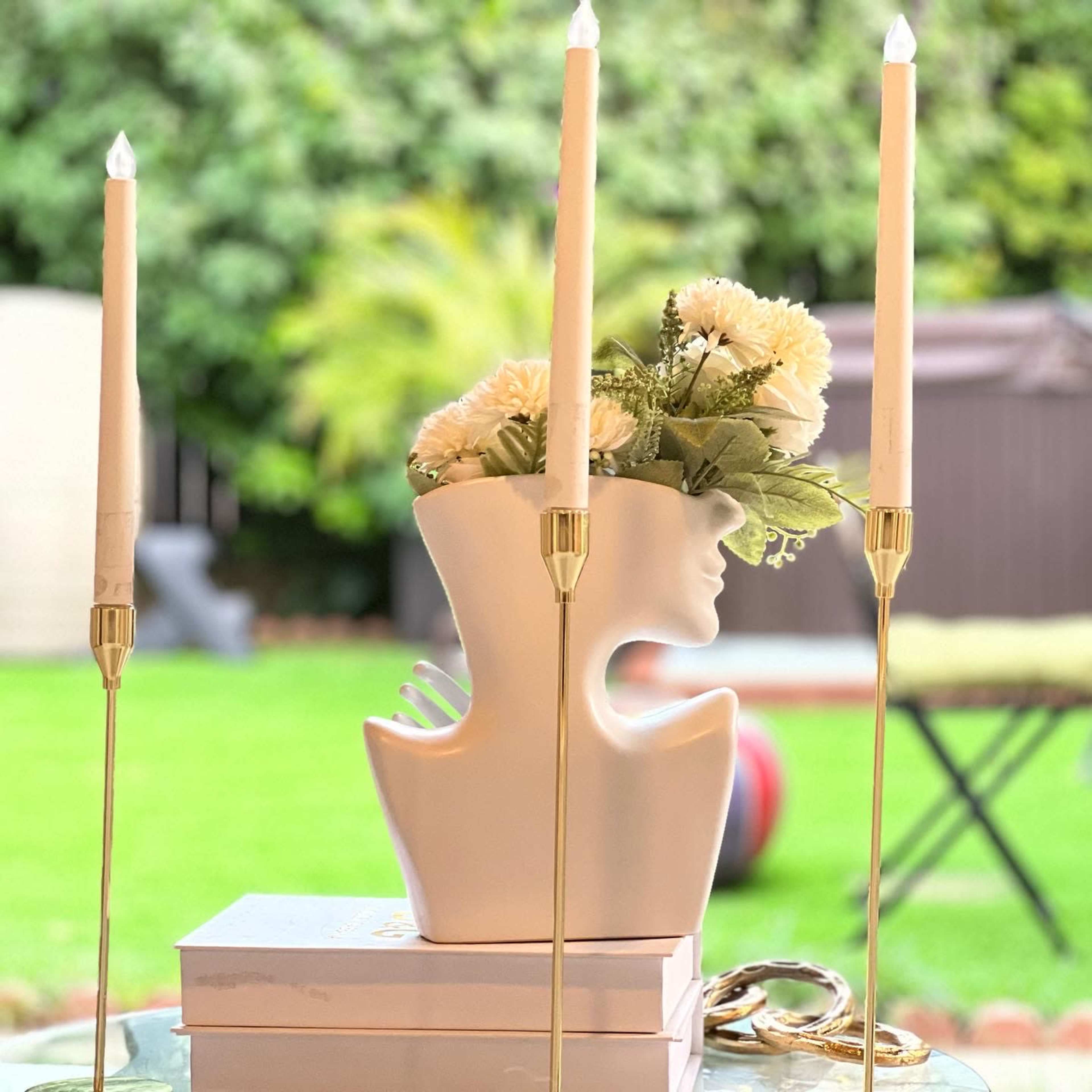 A white vase shaped like a face, holding flowers, flanked by three tall candle holders with taper candles, set against a backdrop of green grass.