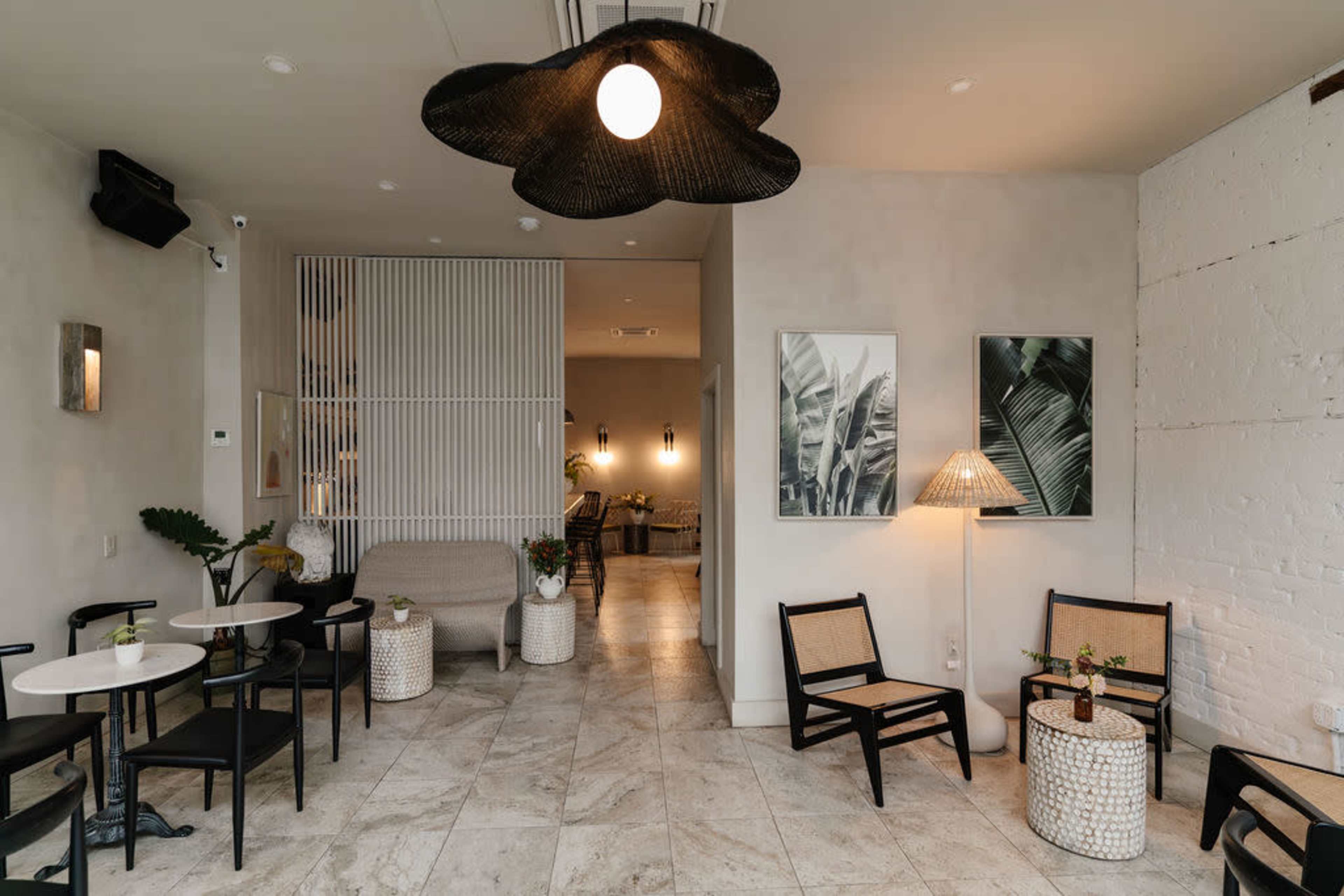 The image shows a stylish, minimalistic interior of a café featuring light-colored walls, woven light fixtures, and a mix of seating options with small tables.