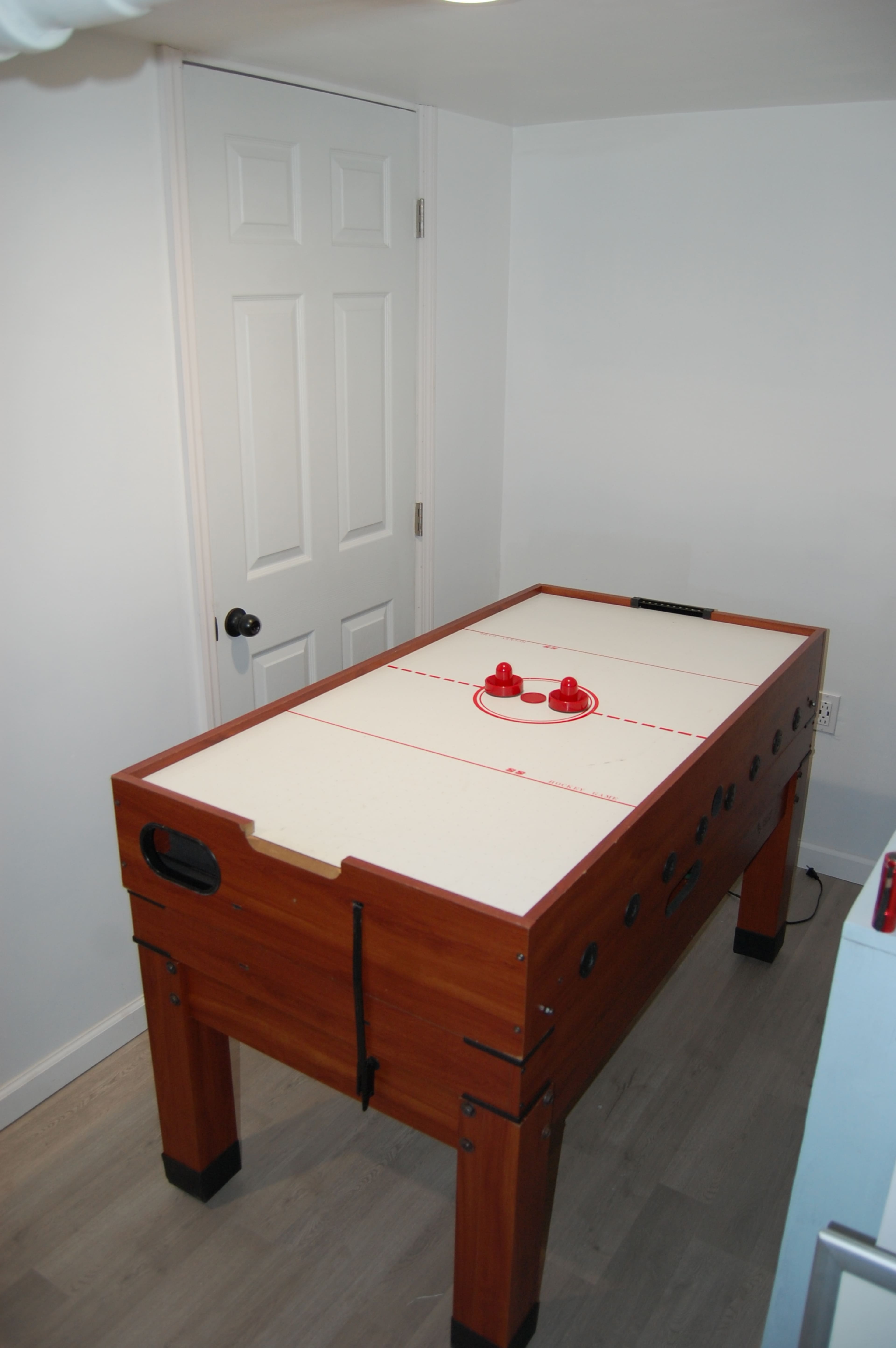 The image shows a wooden air hockey table positioned against a white wall with a closed door.