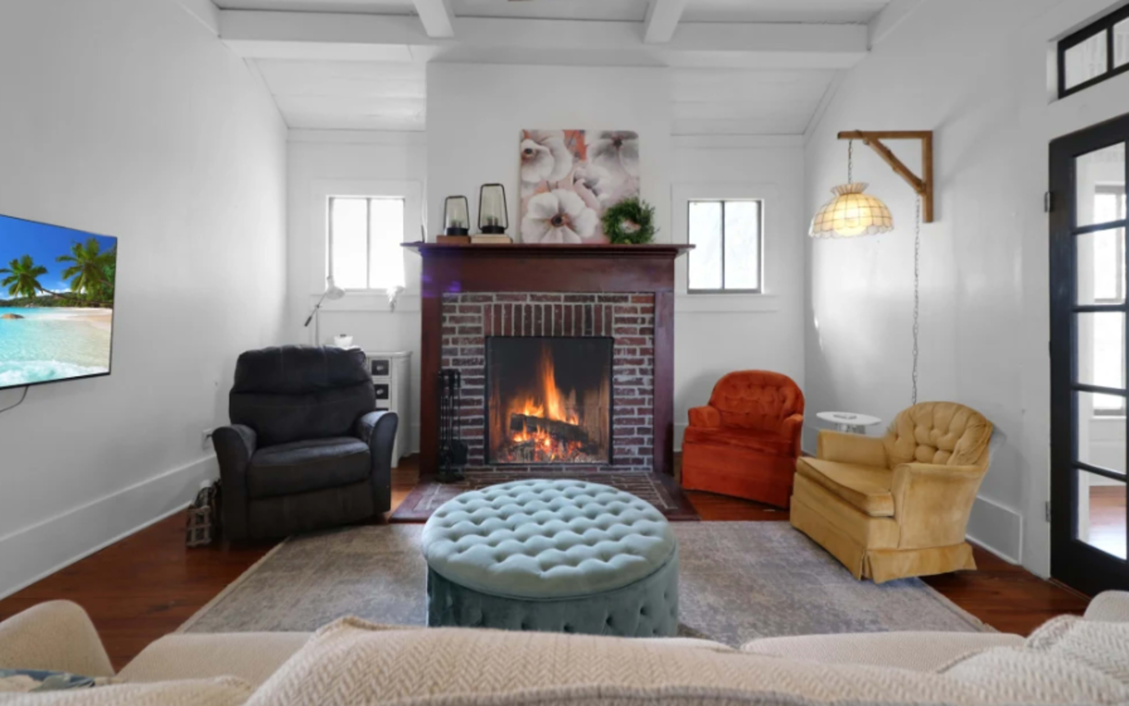 The living room features a brick fireplace with a lit fire, flanked by two armchairs and a tufted ottoman, with a television mounted on the wall opposite.