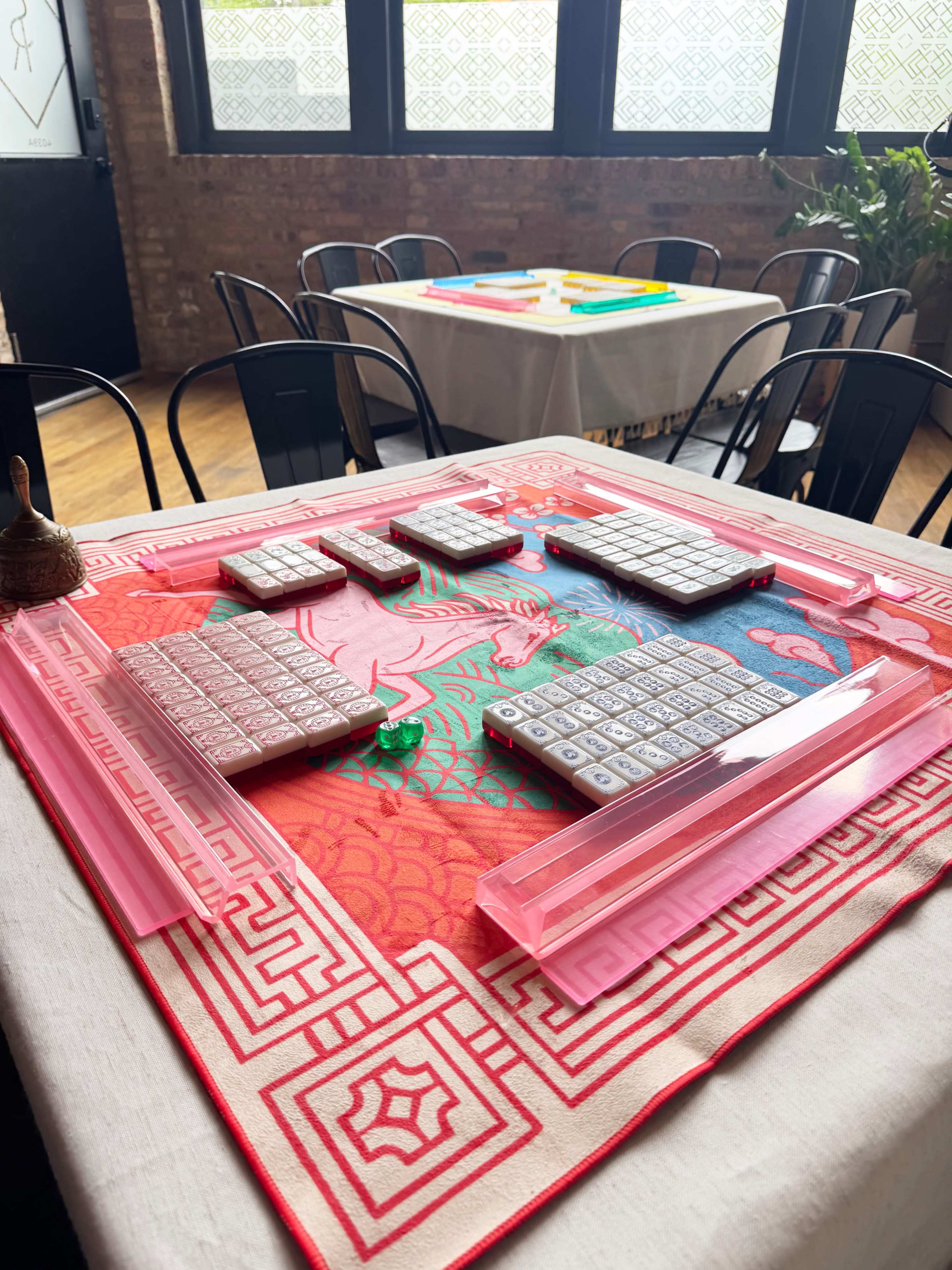 A square table is set with a bright cloth featuring a dragon design, and several Mahjong tiles are arranged on it, along with a pink Mahjong rack and a small bell.
