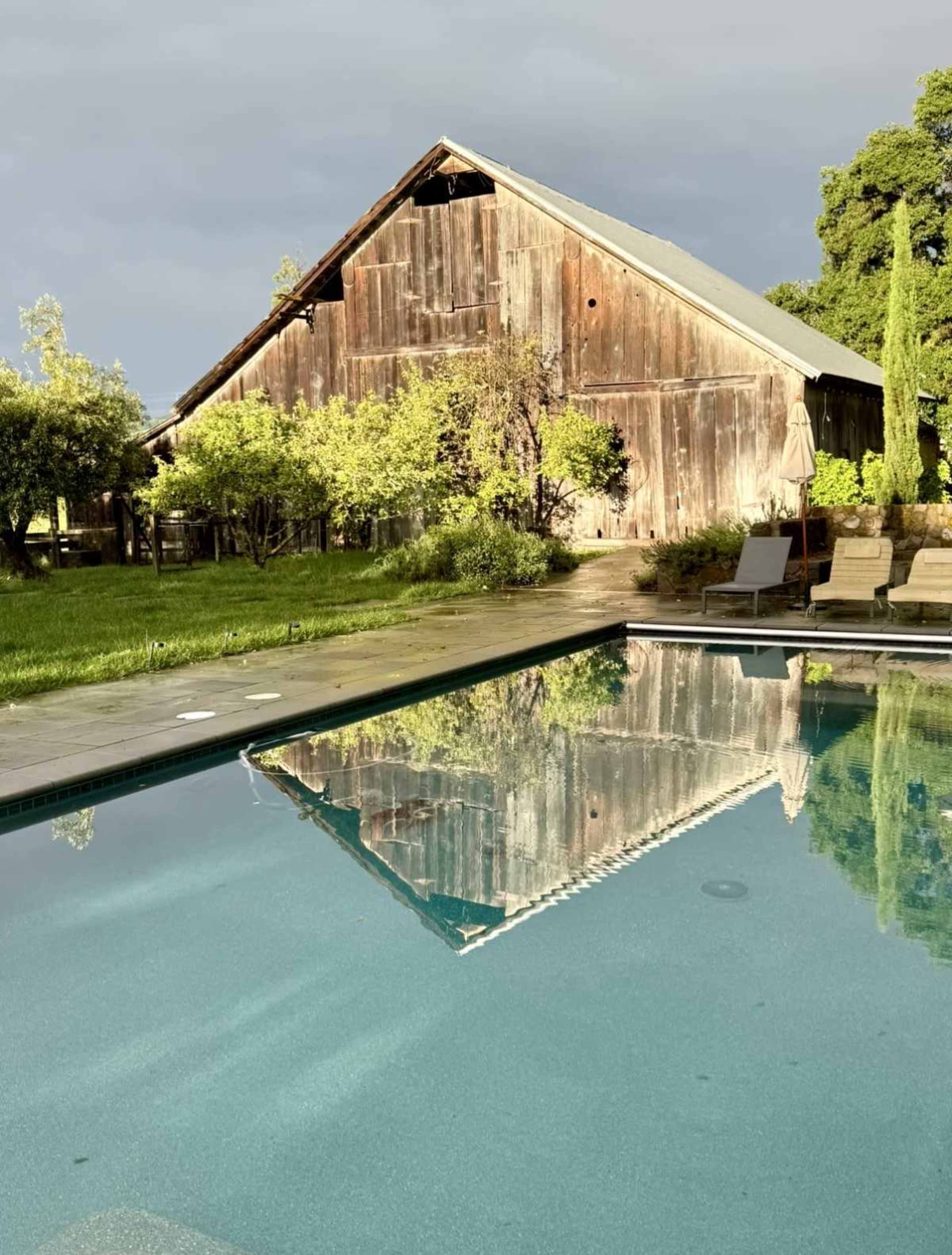 A rustic wooden barn is reflected in the calm water of a swimming pool surrounded by greenery.