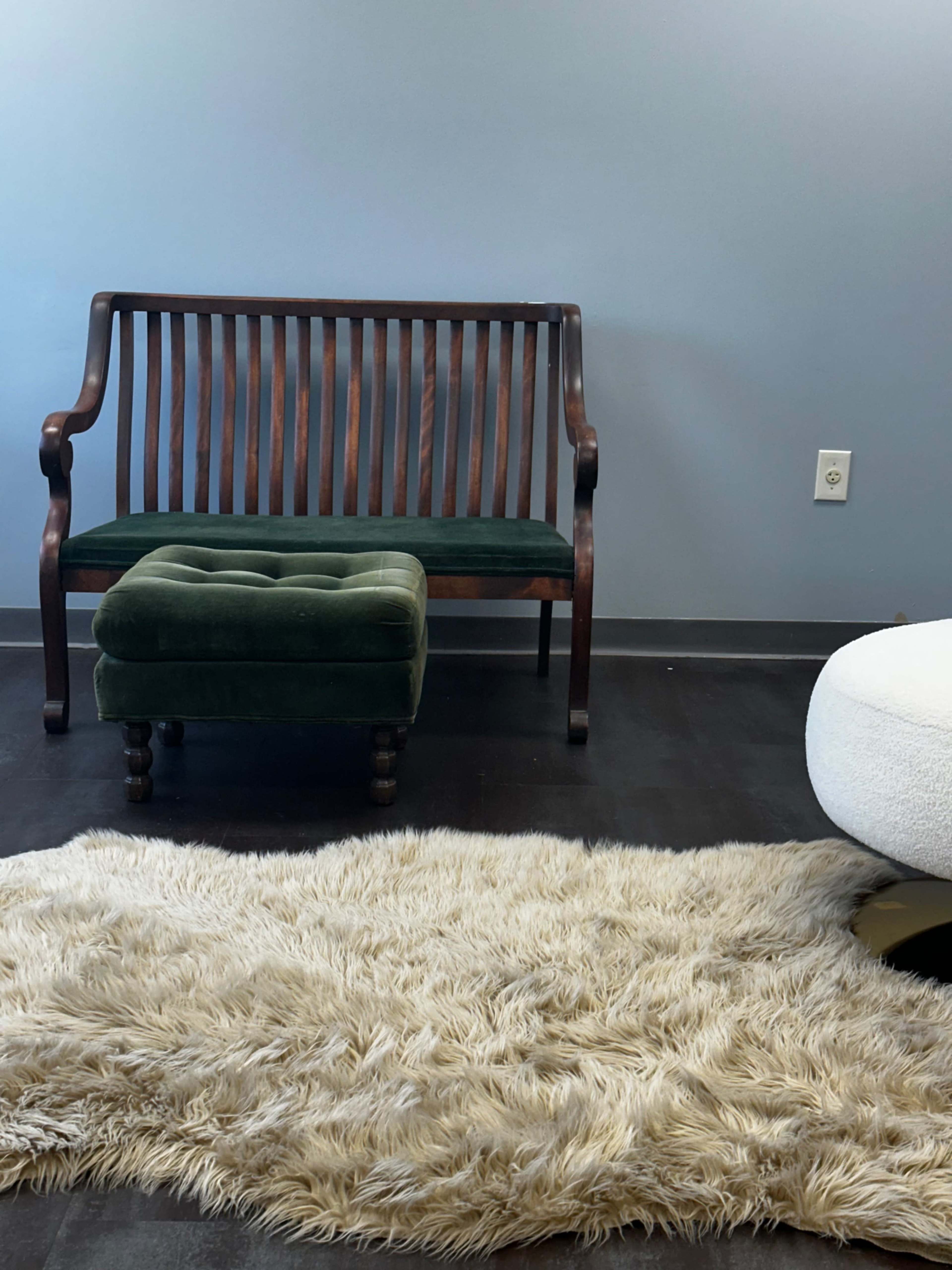 The image shows a wooden bench with a green cushion beside a green ottoman, placed on a fluffy beige rug in a room with light blue walls.