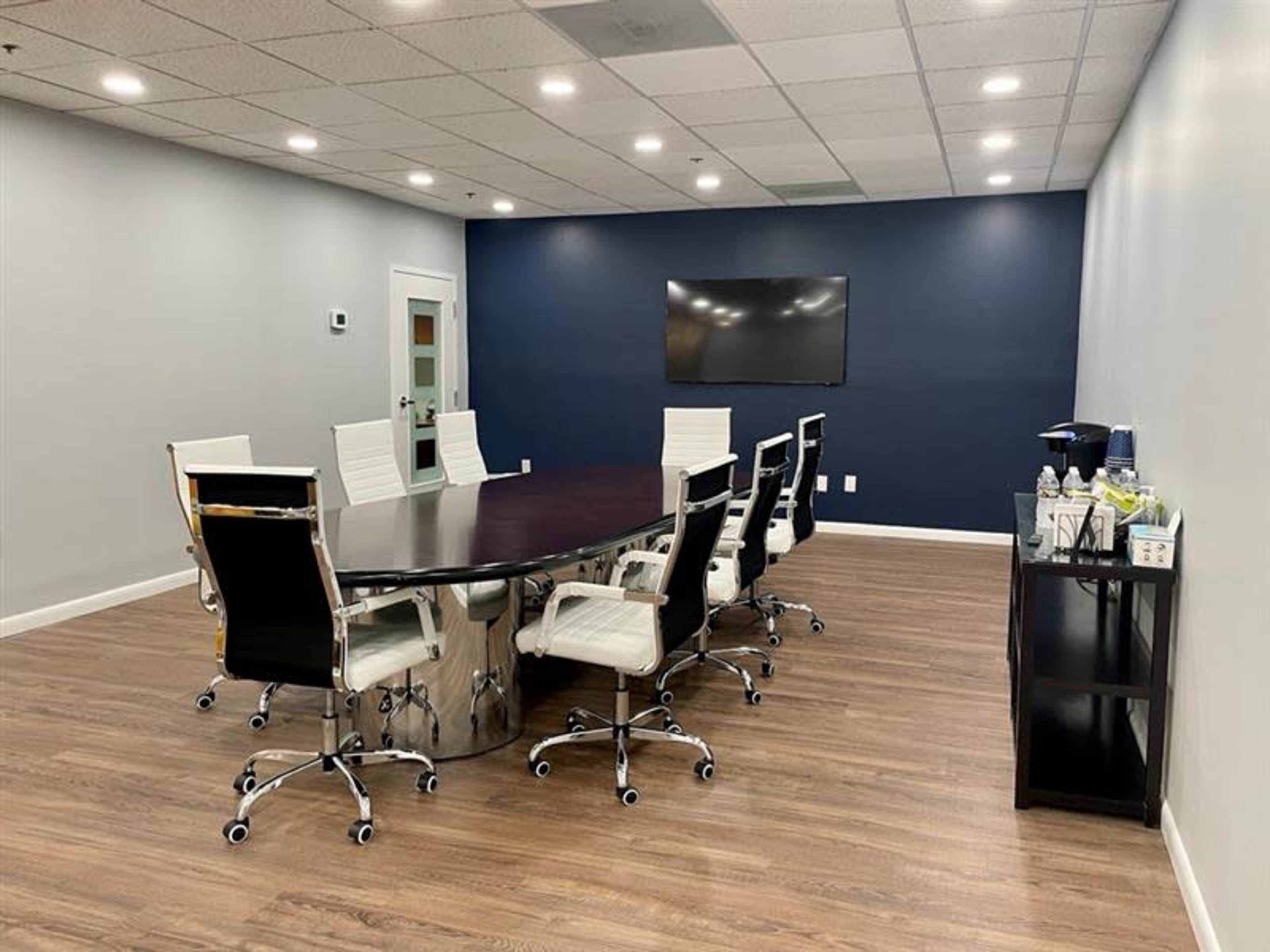 A modern conference room with a large oval table surrounded by eight white and black chairs, a wall-mounted TV, and a coffee station against one wall.