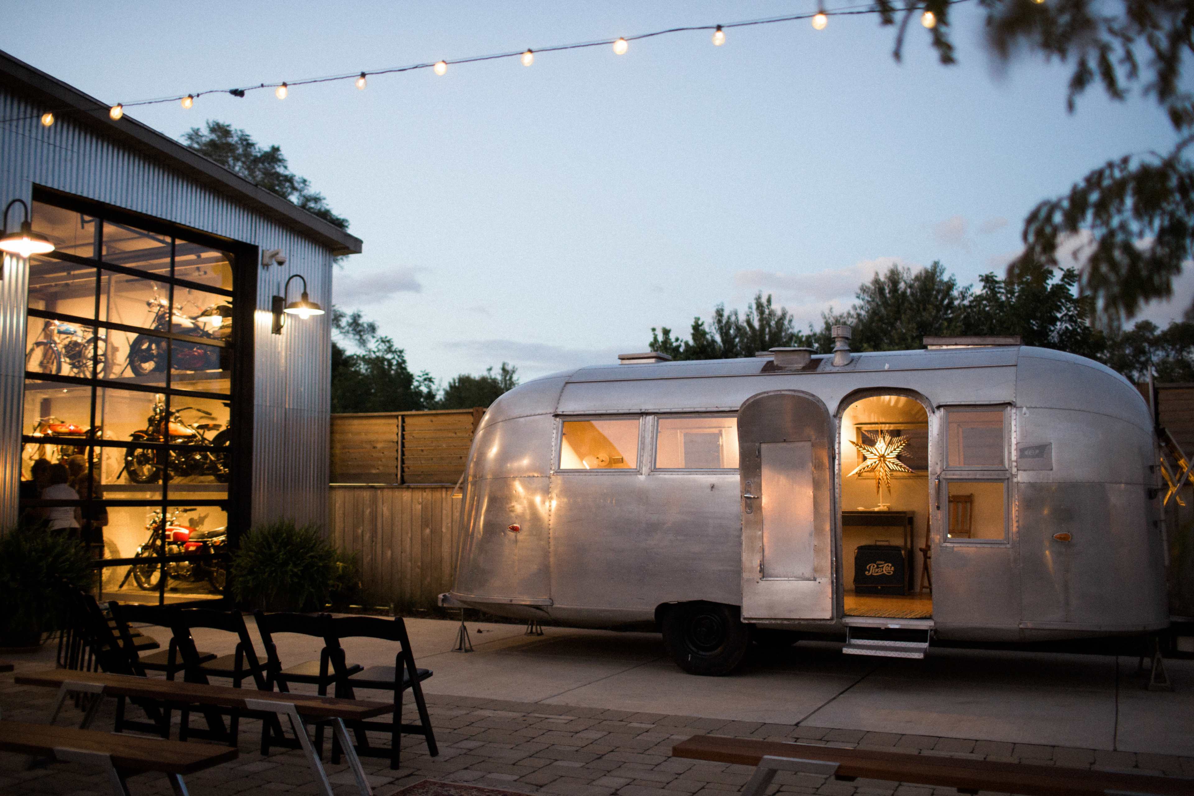 An Airstream trailer is parked outside a modern building with large glass windows, adorned with string lights in the evening.