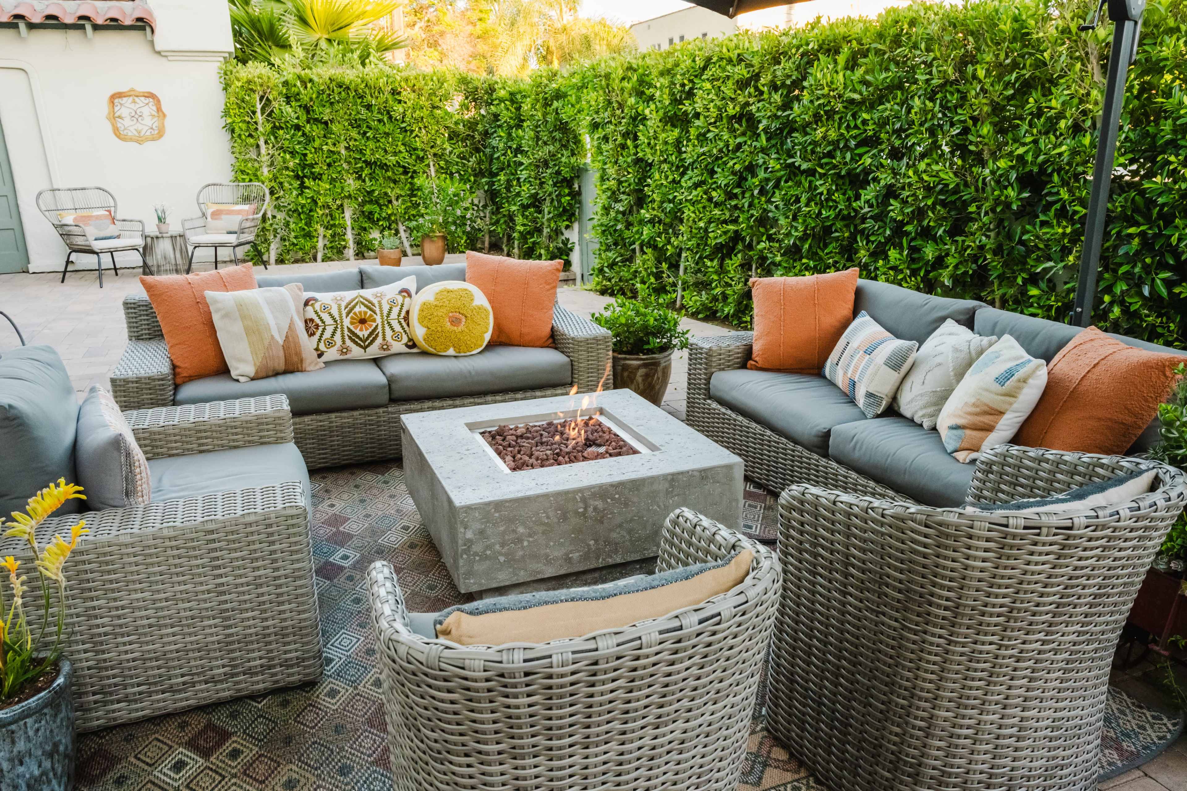 A patio area features a seating arrangement of gray sofas and wicker chairs surrounding a square concrete fire pit.