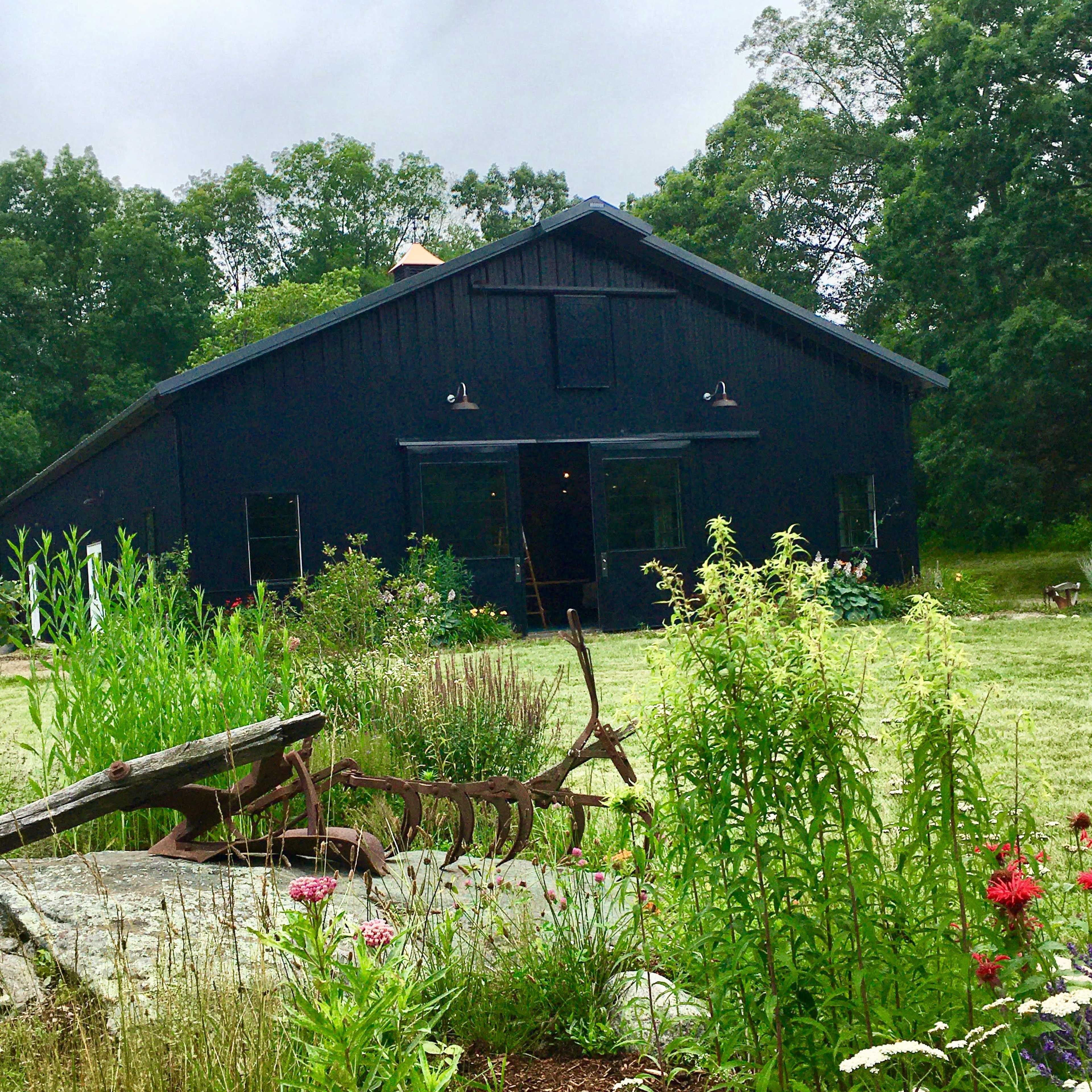 A black barn stands in a rural setting surrounded by green plants and flowers.
