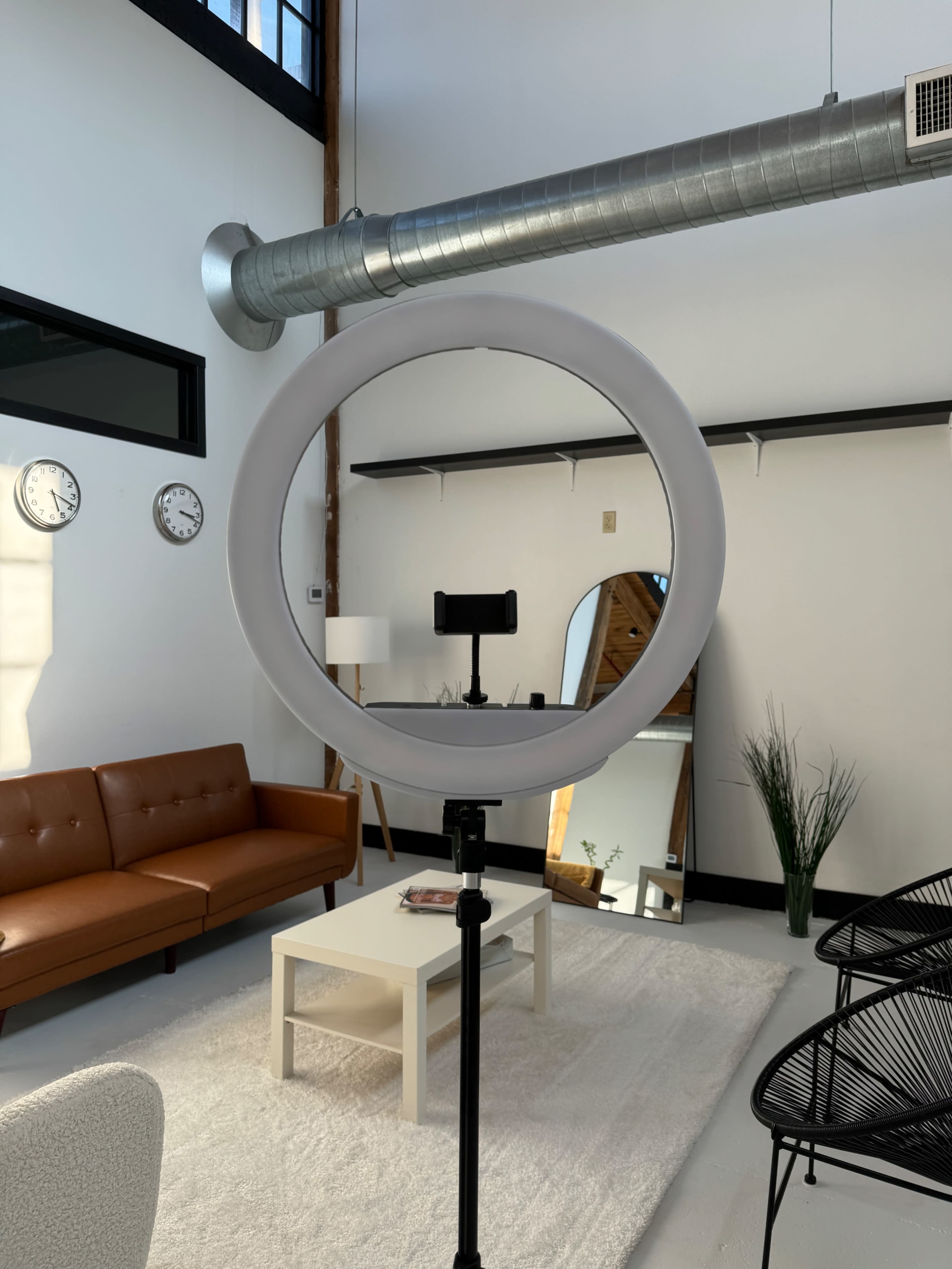 A circular ring light is set up in a bright, minimalist room featuring a brown sofa, a coffee table, and wall-mounted clocks.