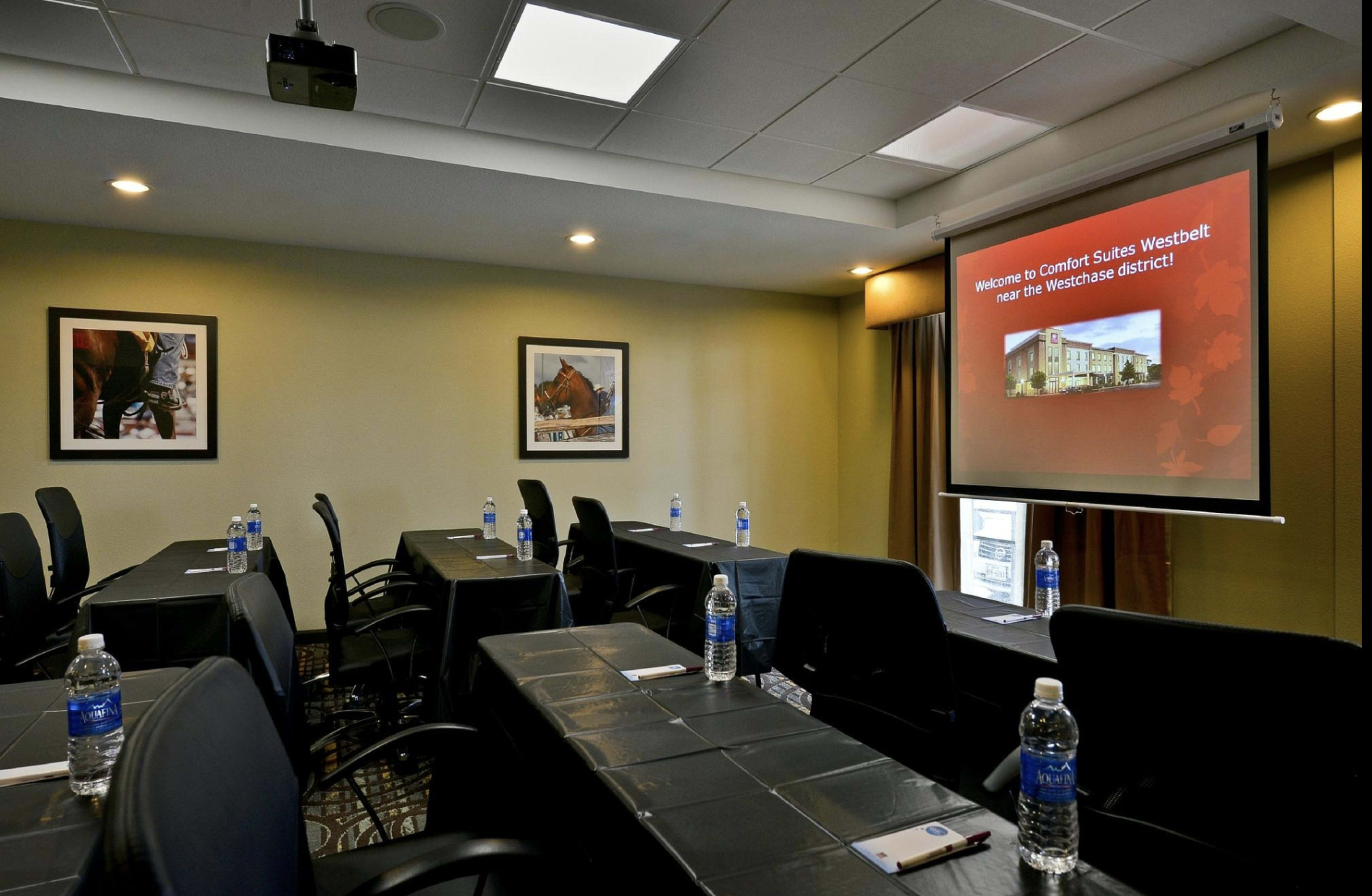 A conference room with multiple black tables and chairs set up, featuring water bottles on each table and a projector screen displaying a welcome message.