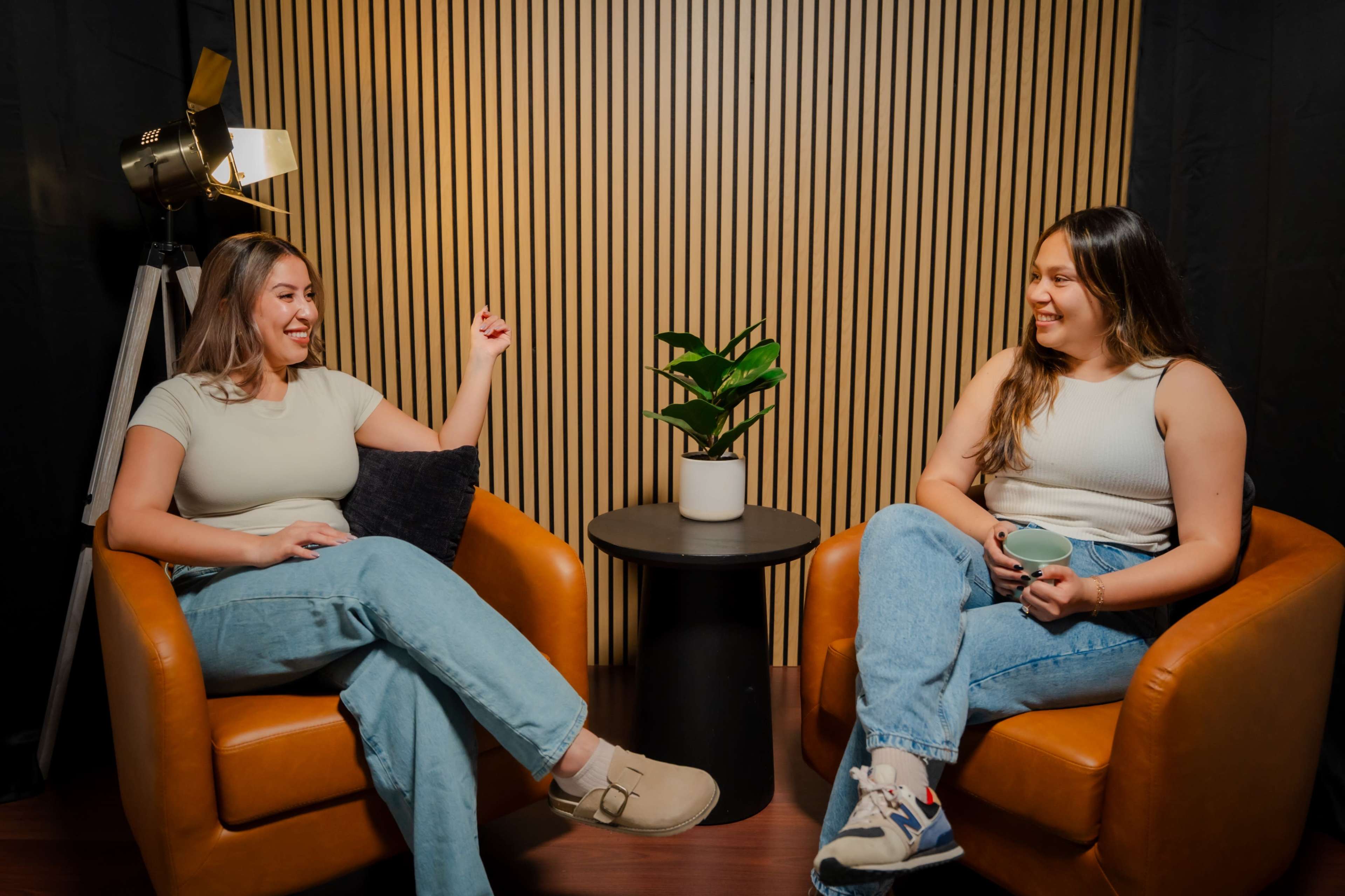 Two women sit in orange armchairs, engaged in conversation, with one gesturing and a plant on a small table between them.