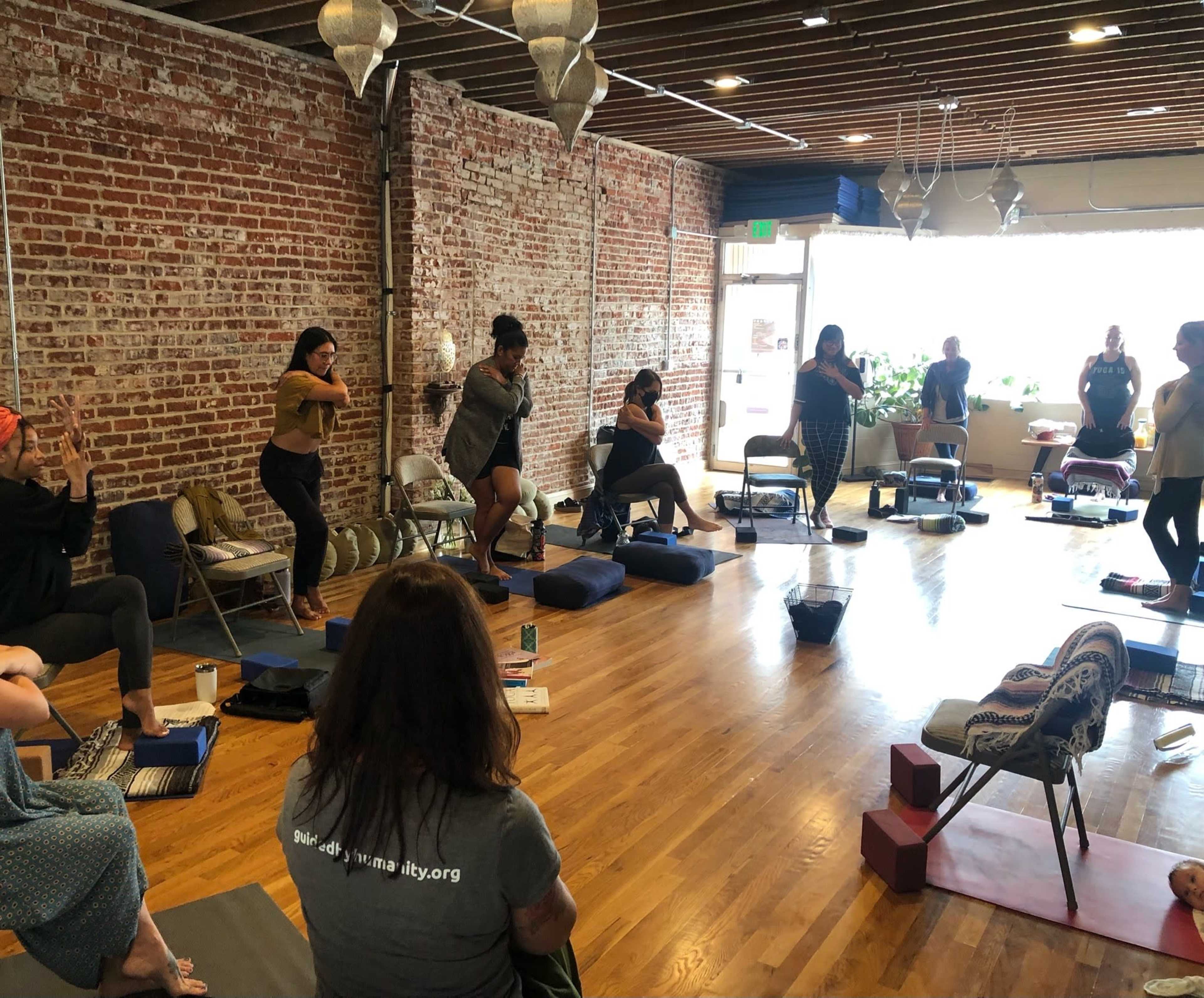 A group of people participates in a yoga class in a spacious studio with exposed brick walls and wooden flooring.