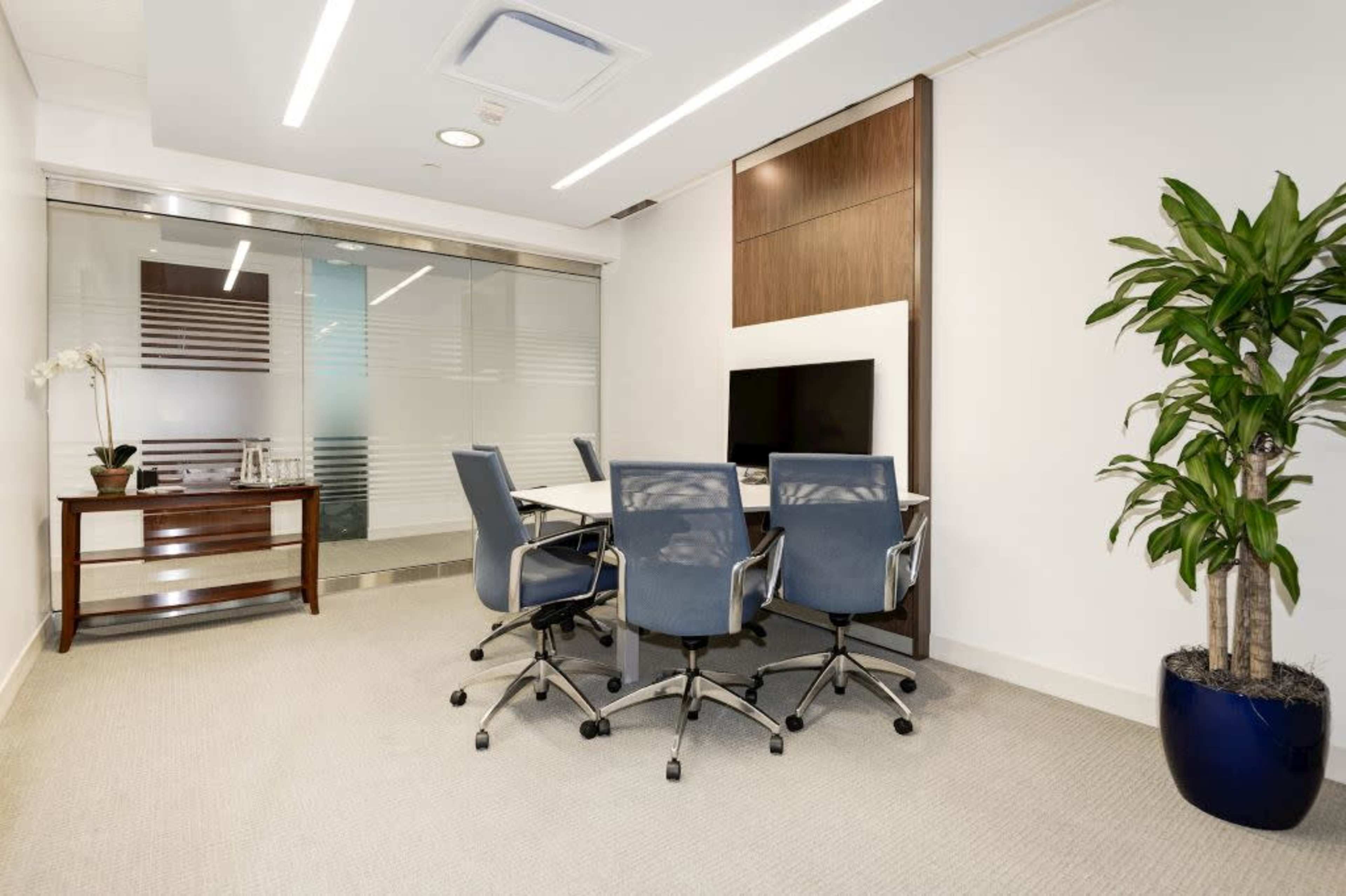 A modern meeting room features a glass wall, a large table with four chairs, a television, and a potted plant.
