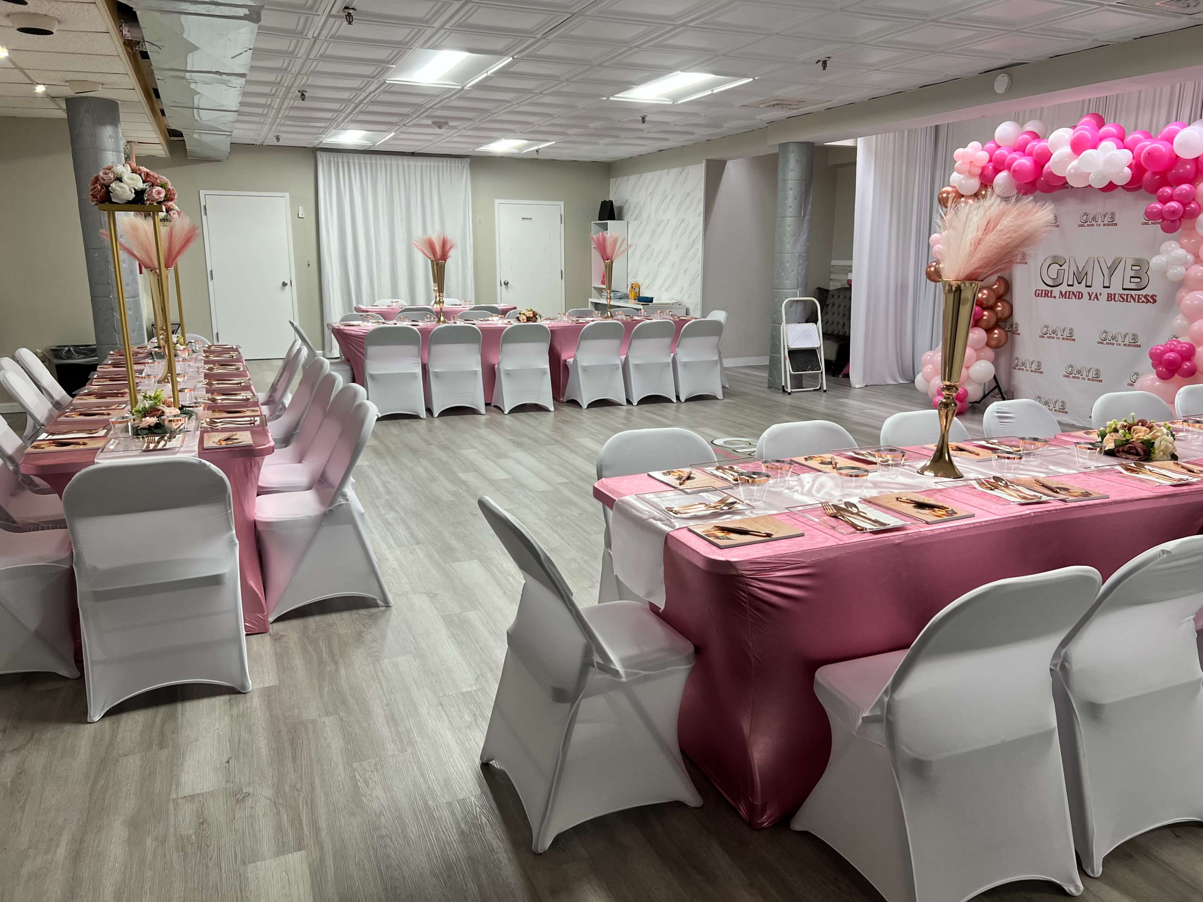 The image shows a decorated banquet hall with tables set in pink and white for an event, featuring floral arrangements and balloon archways.