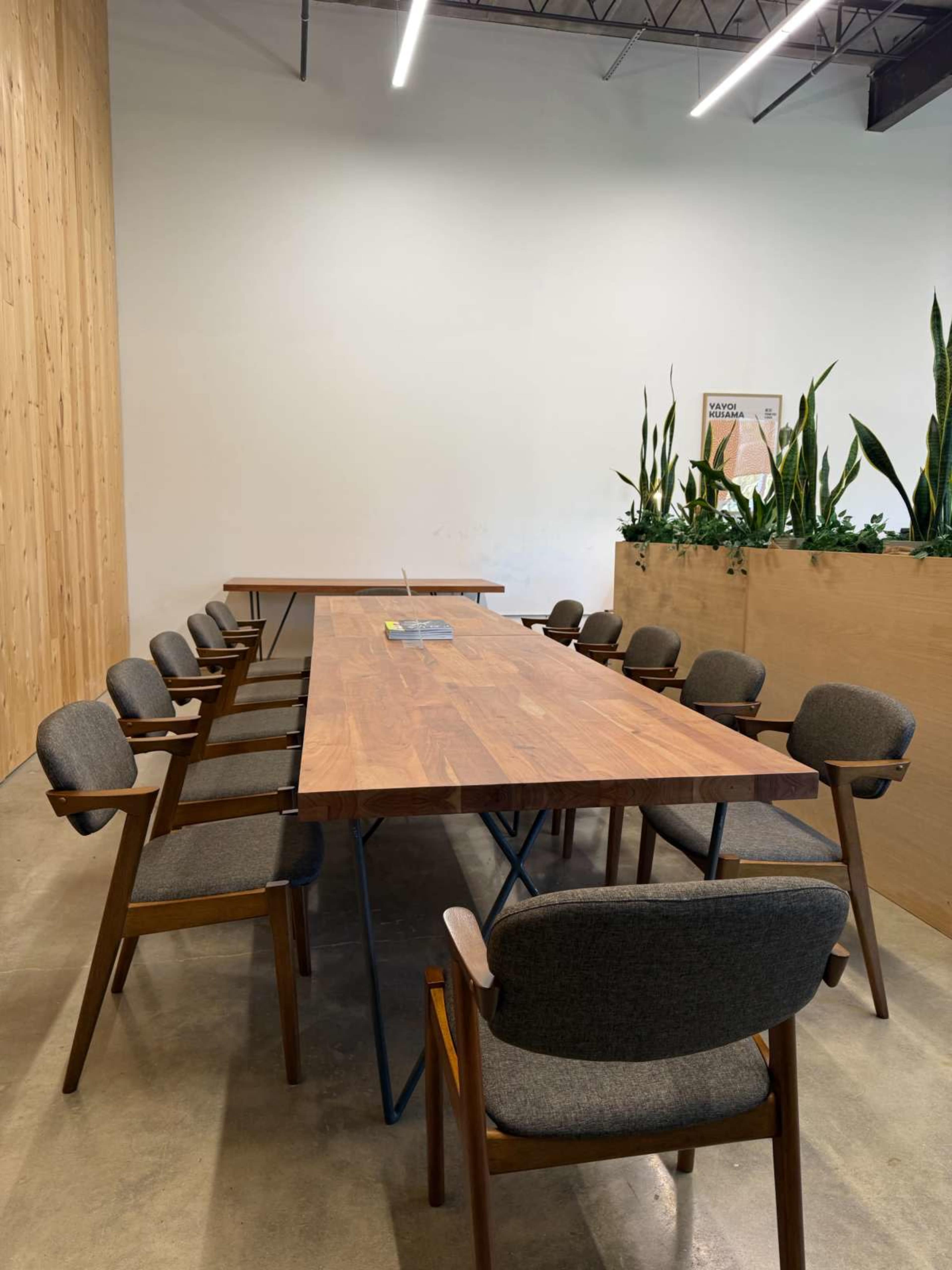 A long wooden table with several chairs is set in a bright room featuring a plant display along one wall.