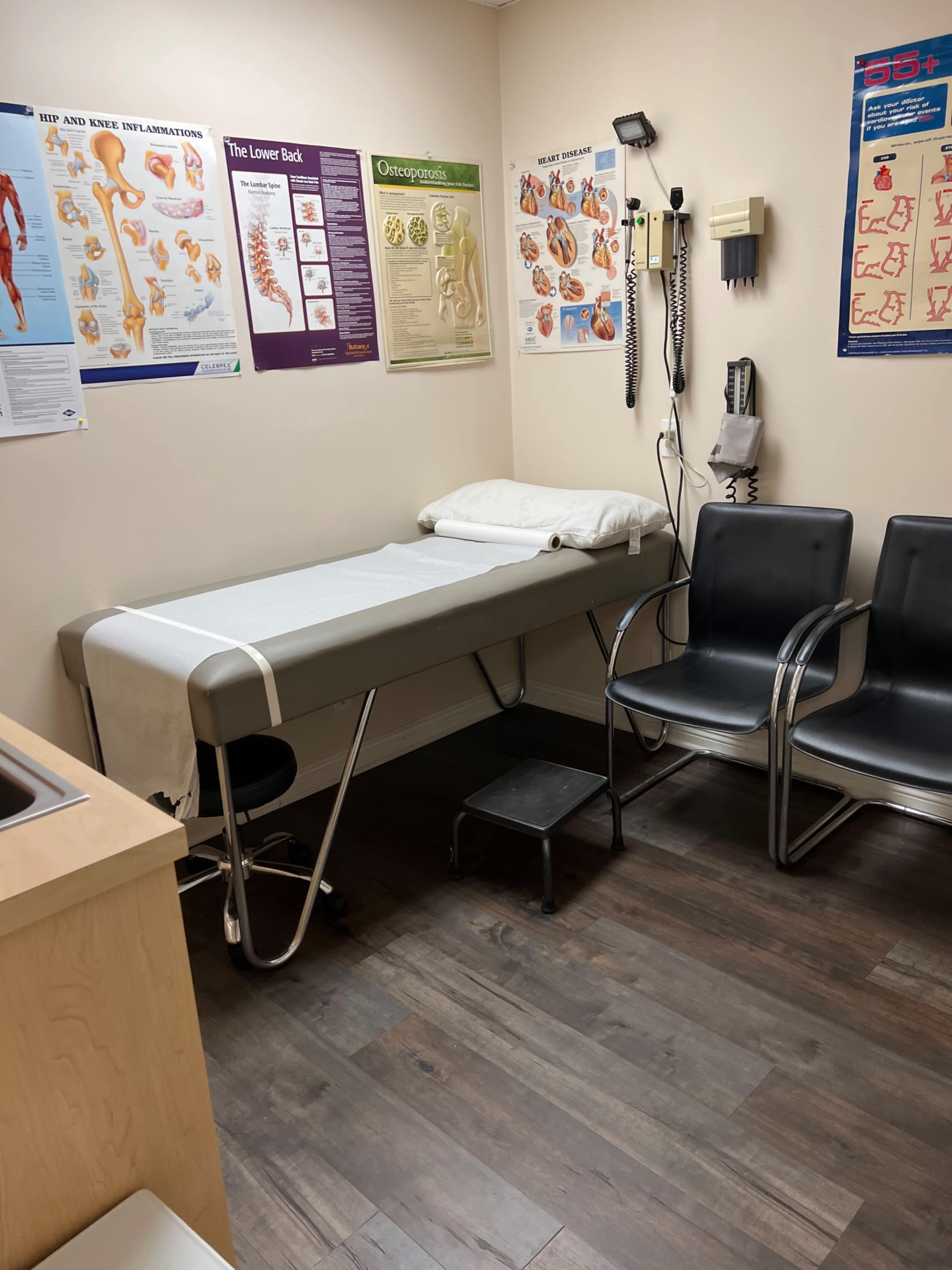 A medical examination room with a treatment table, a stool, and two chairs, along with anatomical posters on the walls.