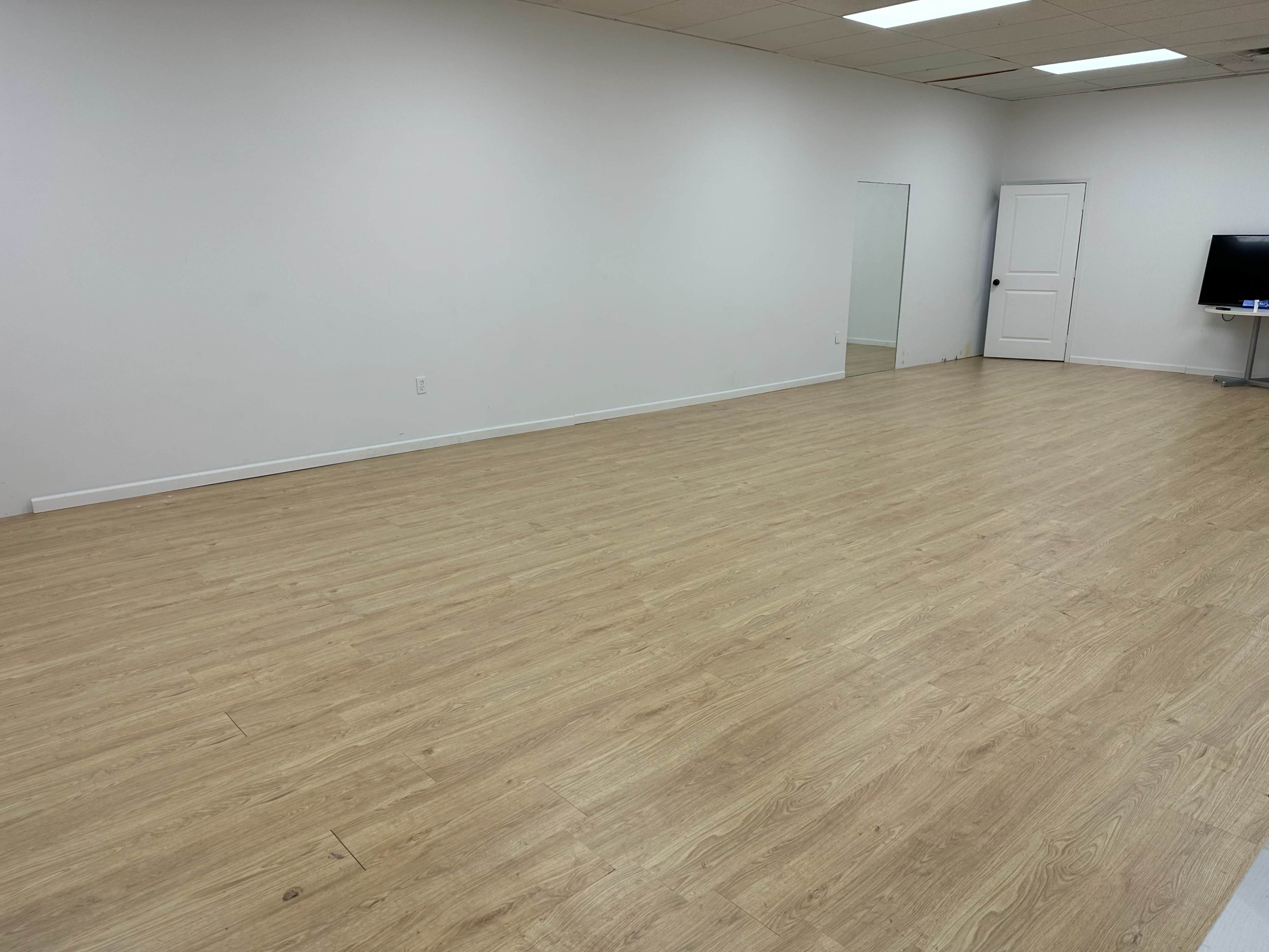 The image shows an empty, well-lit room with wooden flooring, a mirror on one wall, and a closed door in the background.