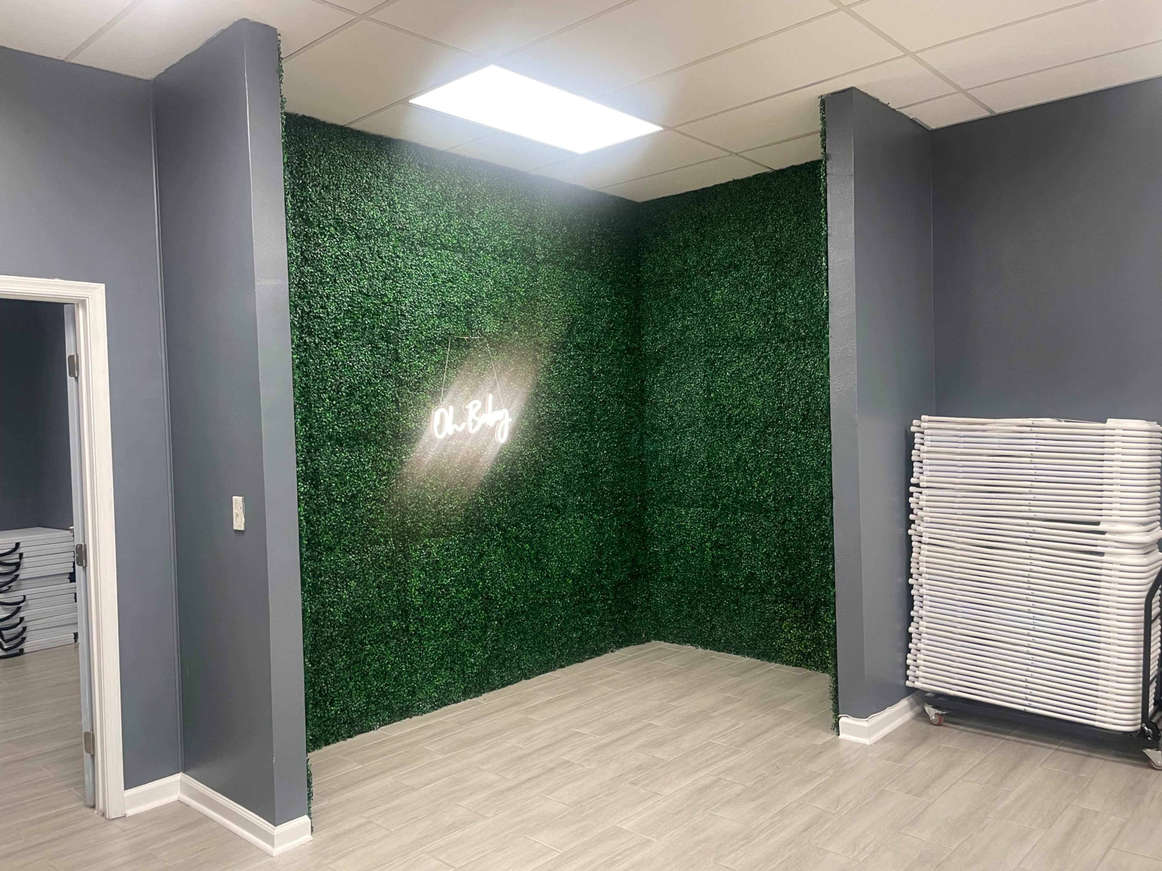The room features a green, artificial grass wall with a glowing sign that reads "Oh, B." and has light-colored walls and a stack of white chairs in the corner.