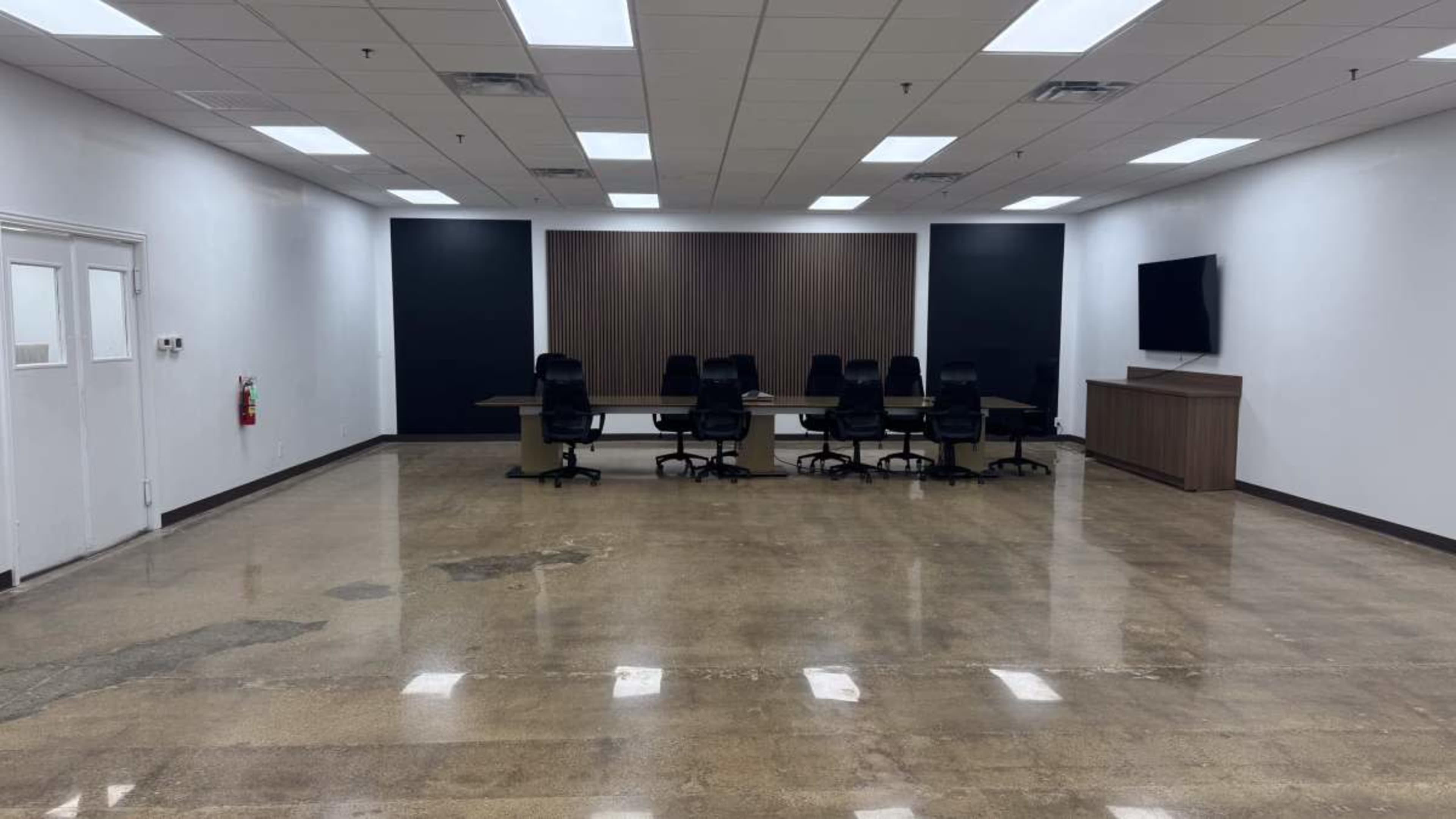 The image shows a large, empty conference room featuring a long table surrounded by chairs, with a wooden accent wall and a television mounted on one side.
